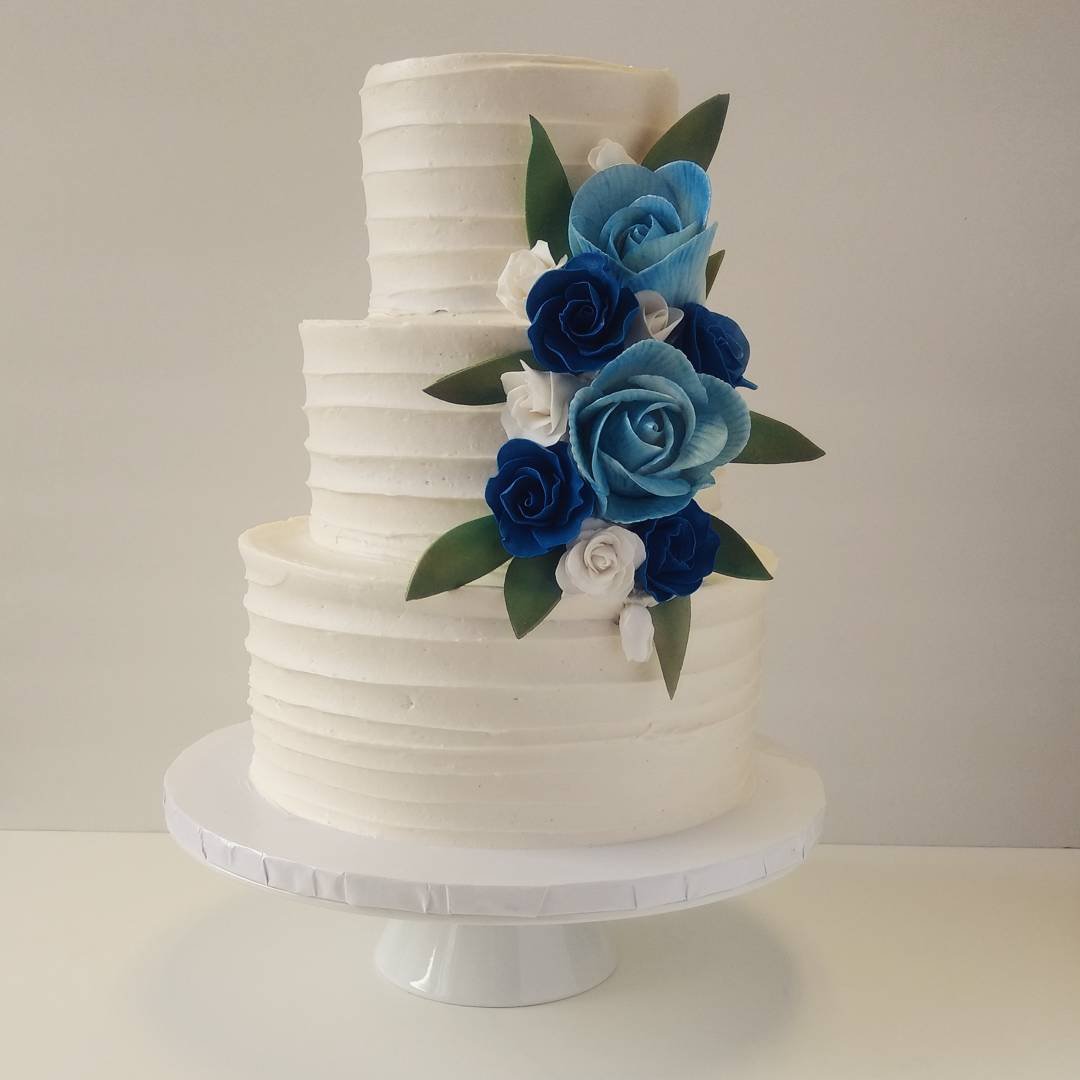 A three-tier wedding cake with a textured white finish, adorned with blue and white roses and green leaves