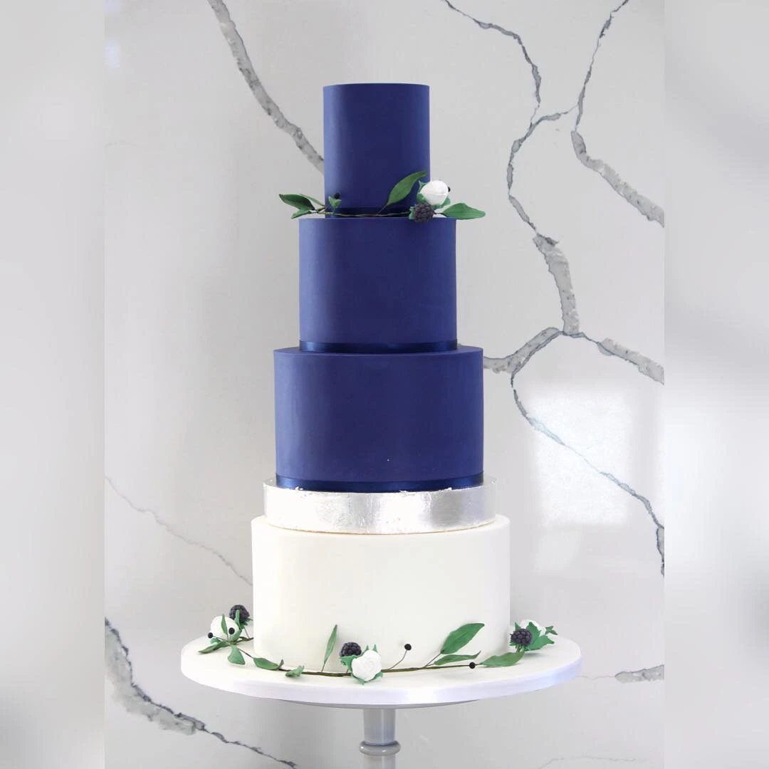 A four-tier wedding cake featuring deep blue and white layers, adorned with silver accents and fresh greenery