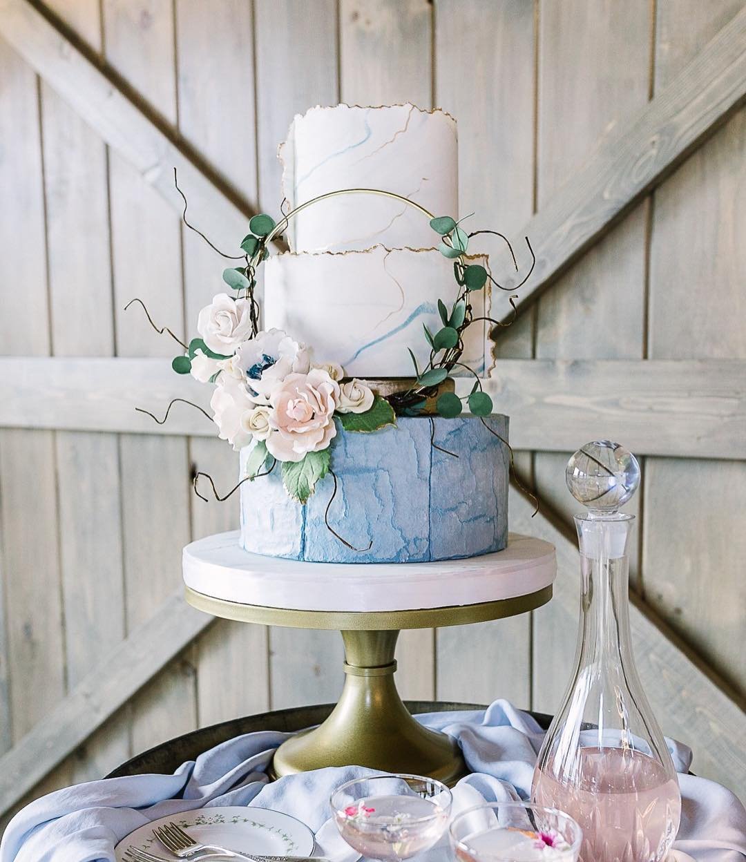 A three-tier wedding cake with a marbled blue base and white layers, decorated with flowers and greenery, presented on a decorative stand.