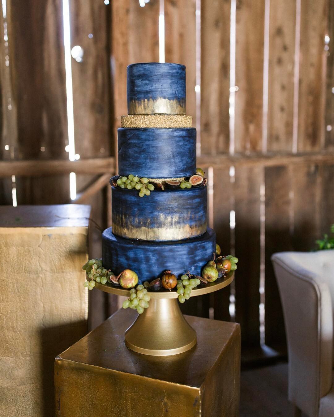 A four-tier cake with navy blue and gold layers, decorated with grapes and figs.