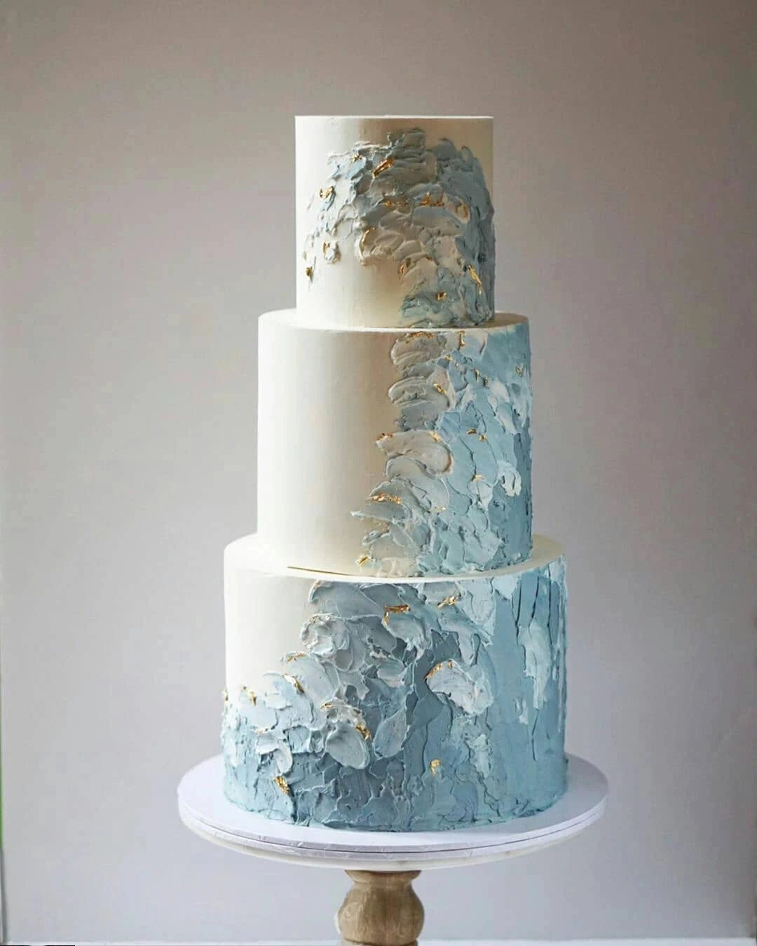 A three-tier wedding cake featuring an abstract blue and white design with gold accents, presented on a simple cake stand.