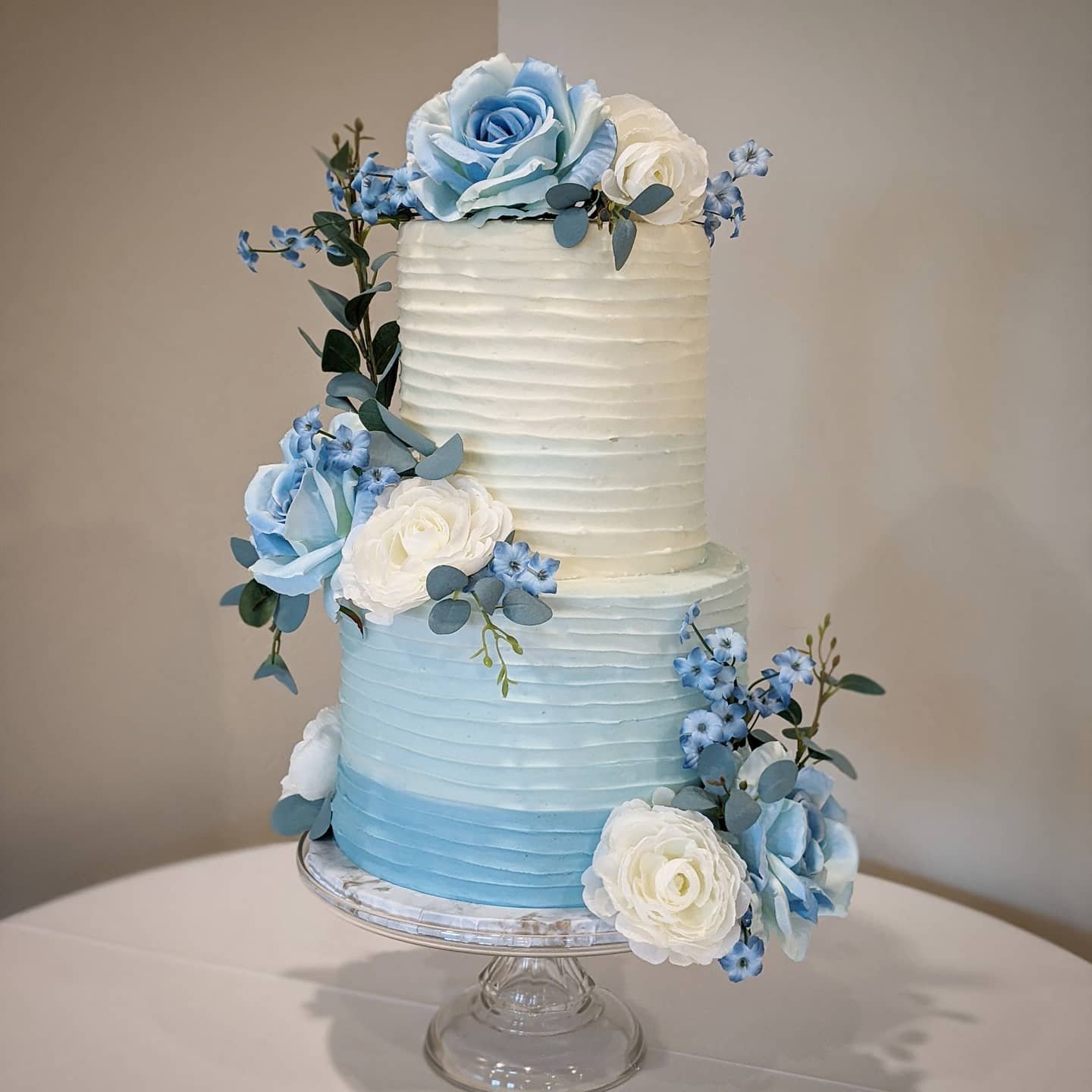 A two-tier wedding cake with an ombre design from cream to soft blue, decorated with blue and white flowers and greenery, displayed on a glass stand.