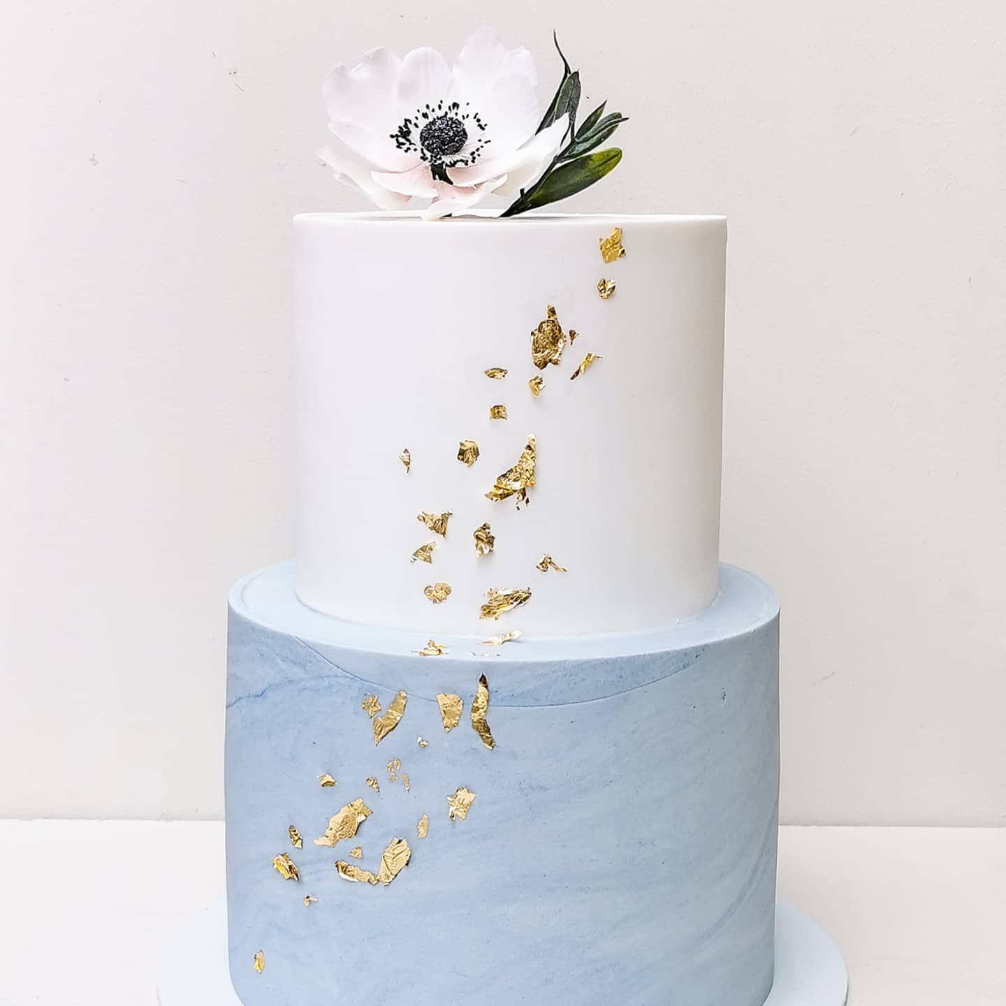 A two-tier wedding cake with a white upper layer and a marbled blue lower layer, adorned with gold leaf accents and topped with a single flower.