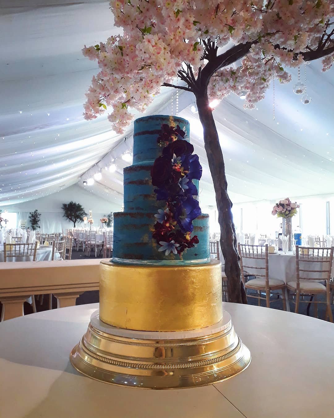 A three-tier cake with blue and gold, decorated with vibrant flowers.