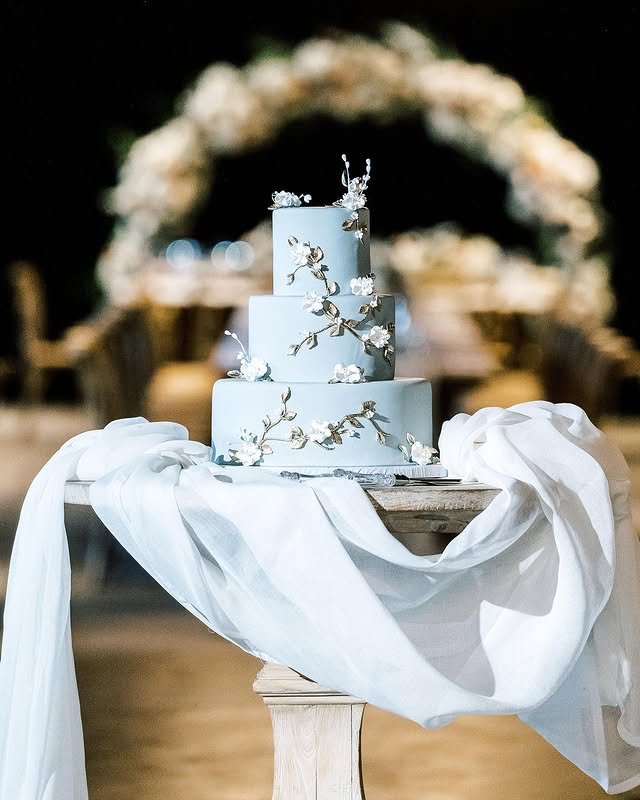 Three-tiered blue wedding cake decorated with sugar flowers and branches, set on a draped table.