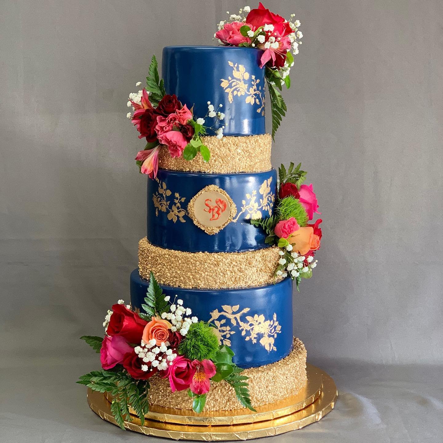 A four-tier cake with navy blue and gold, decorated with colorful flowers and a monogram.