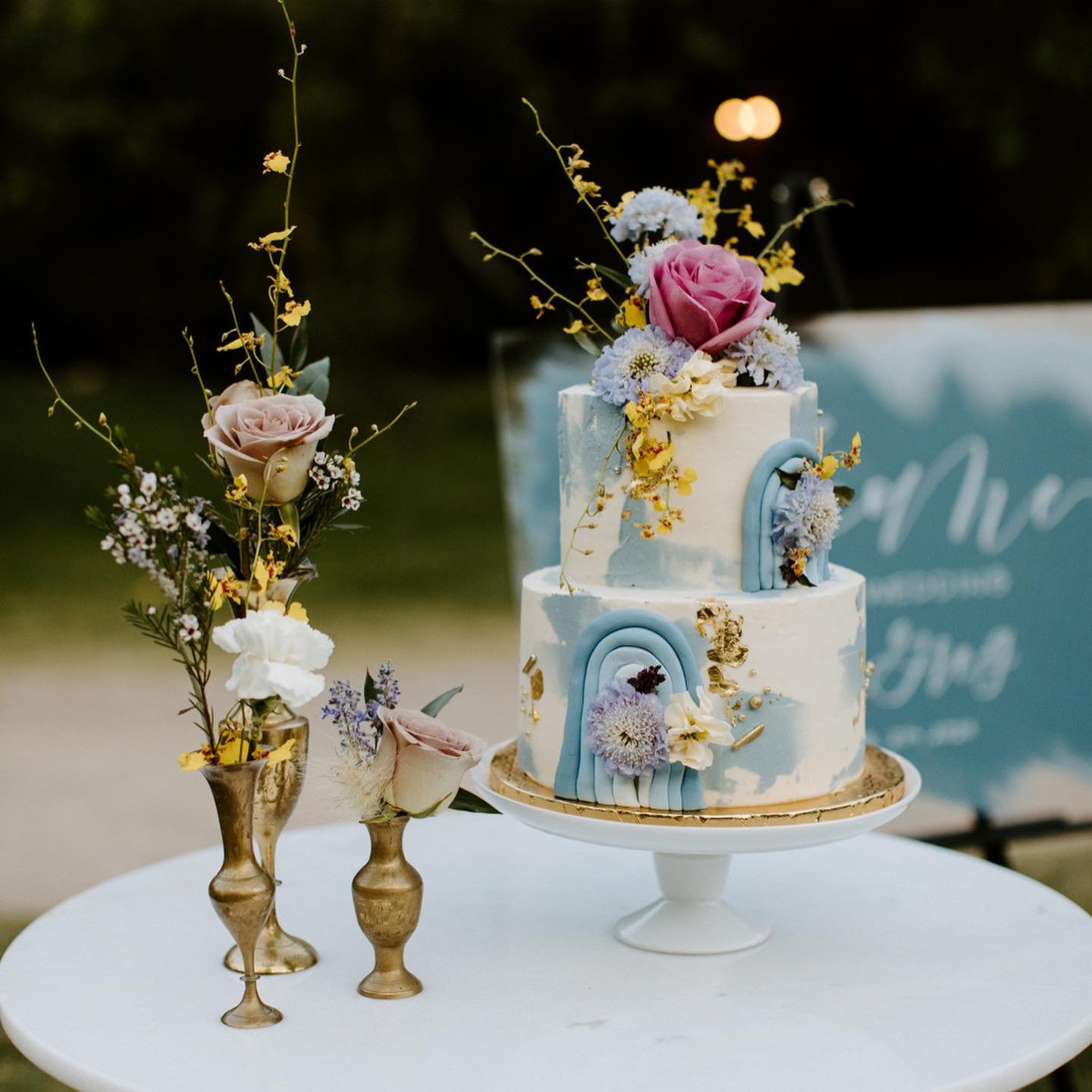 A two-tier wedding cake with white and blue arches, decorated with colorful flowers and gold accents, placed on a round table next to elegant vases of flowers.