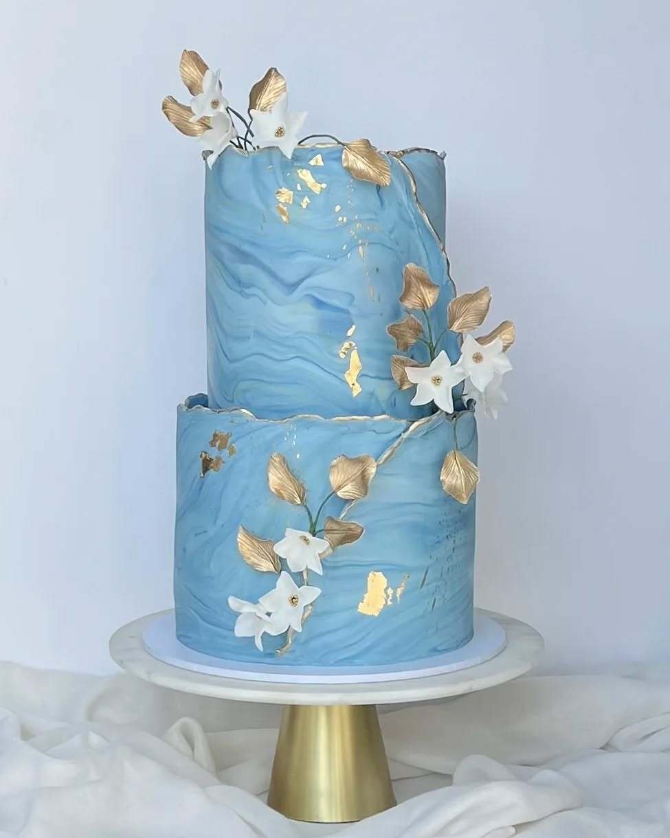 Two-tiered marbled blue wedding cake adorned with gold leaf and white flowers.