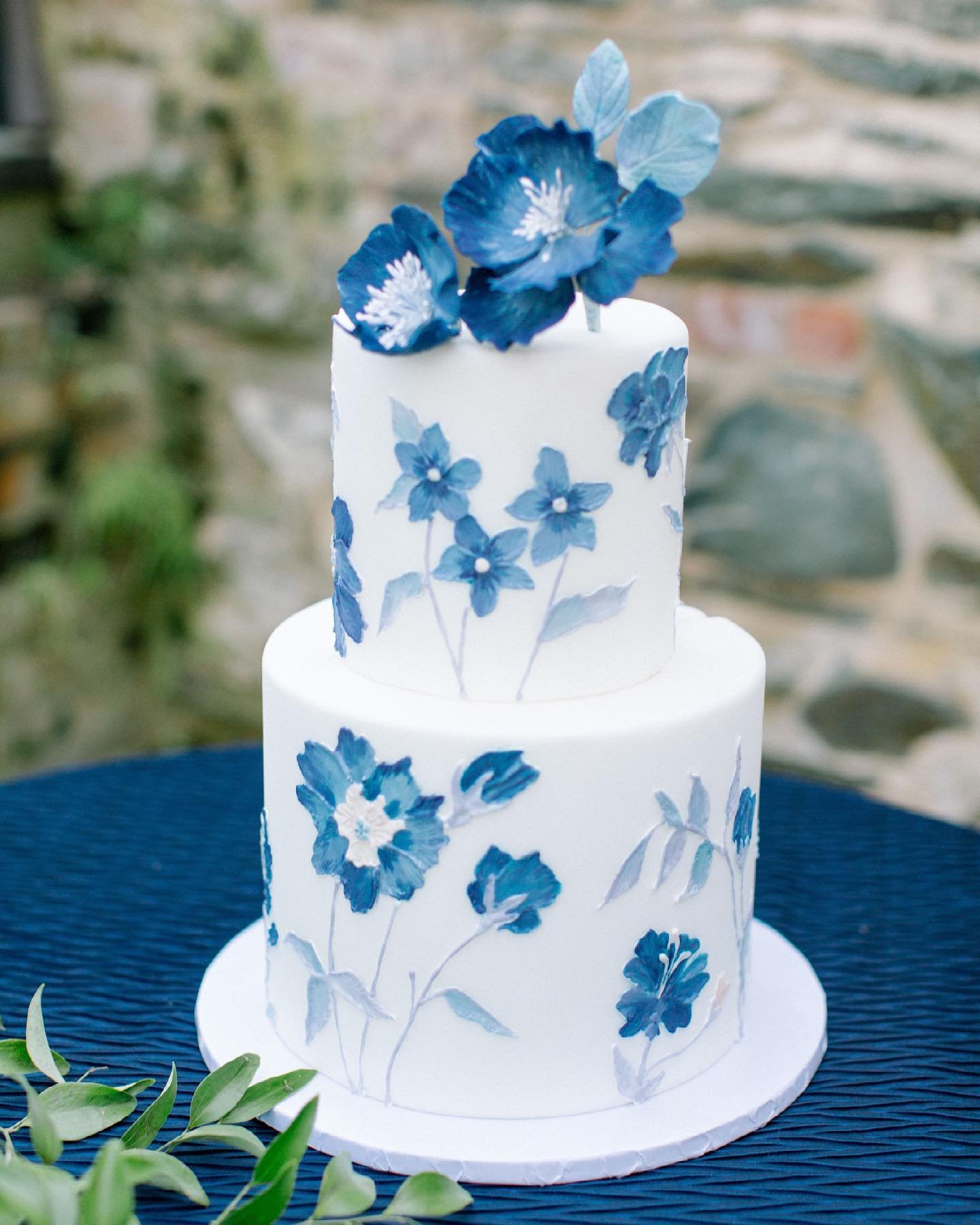 A two-tier wedding cake featuring hand-painted blue floral designs on a white background, elegantly displayed on a table with greenery.