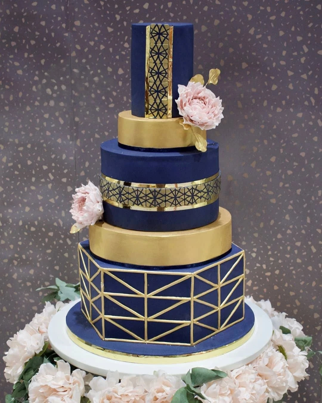 A four-tier cake with navy blue and gold, decorated with pink peonies and geometric patterns.