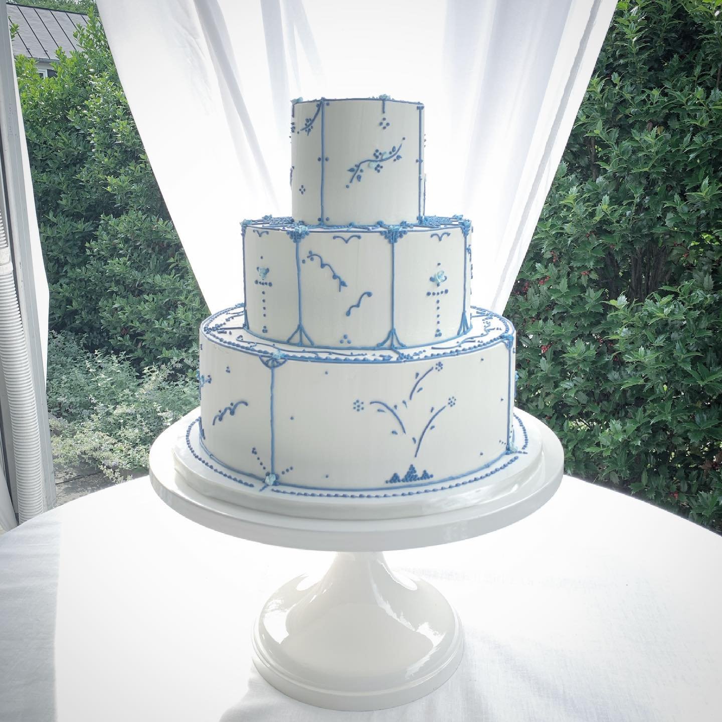 A three-tier wedding cake with white icing and detailed blue decorations, displayed on a white stand in an outdoor setting surrounded by greenery.