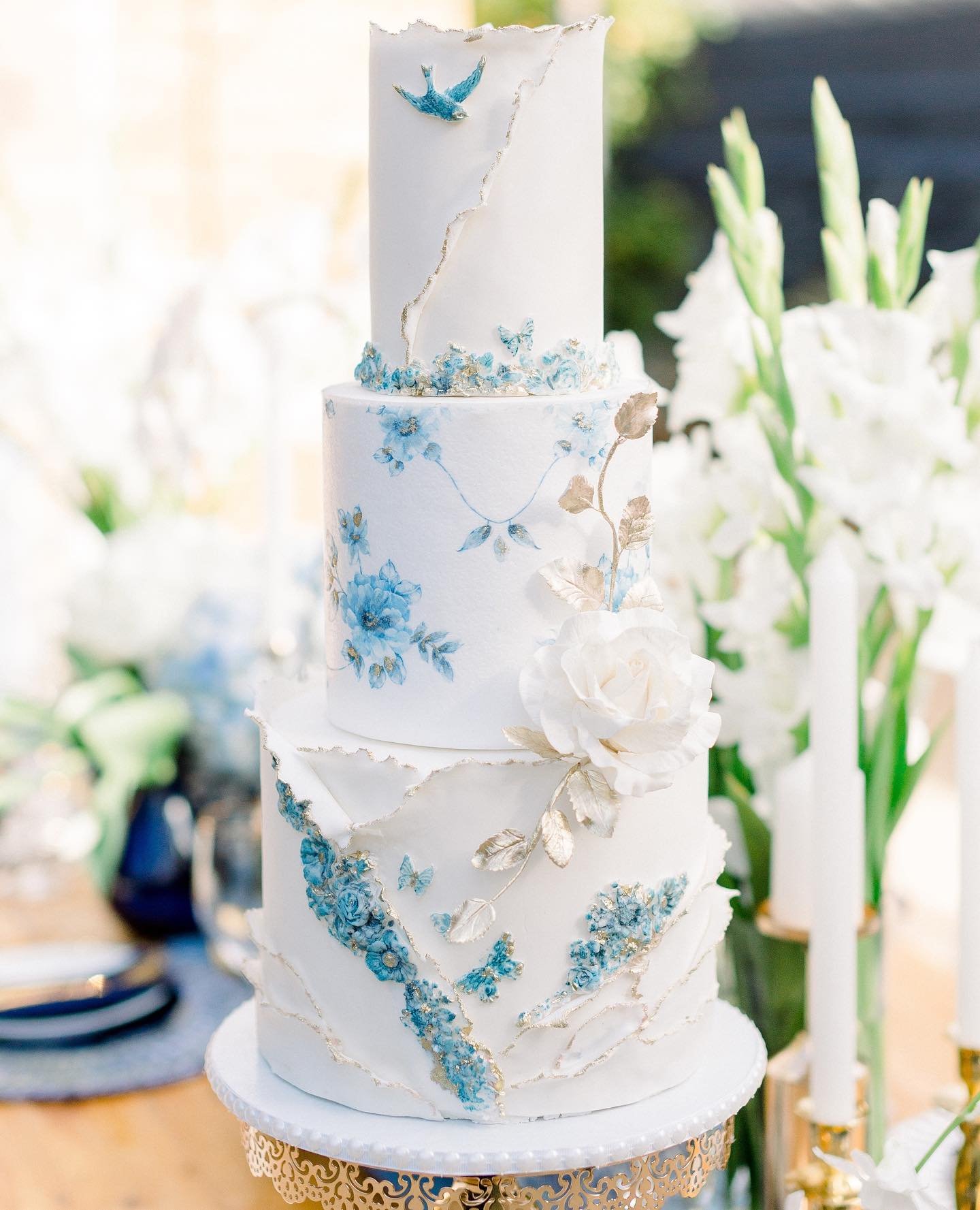 A three-tier wedding cake with white icing, blue floral decorations, and gold accents, beautifully displayed on a decorative stand amidst an elegant table setting.