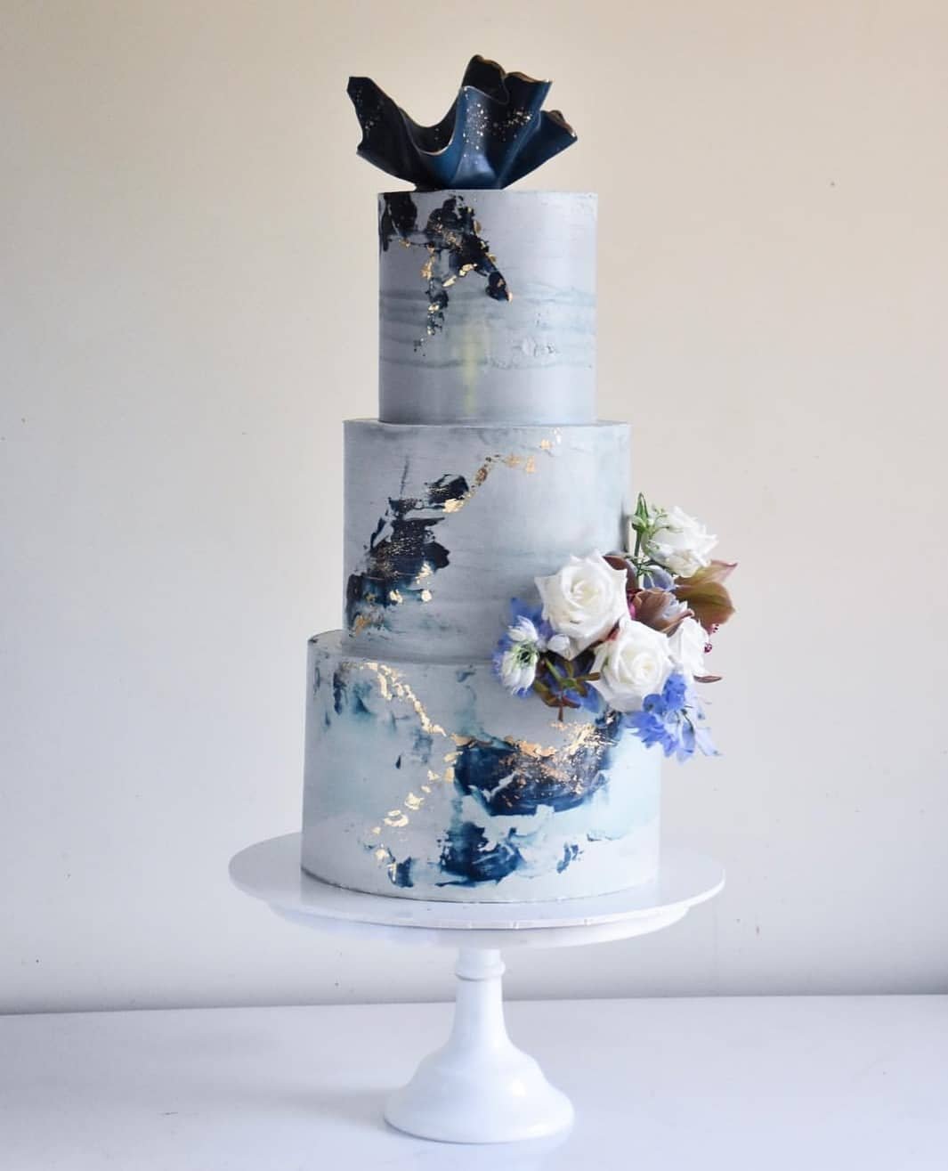 A mesmerizing light blue wedding cake with three tiers, each adorned with intricate floral patterns. The top tier showcases delicate sugar roses and white blooms, while the bottom tier features elegant lace-like piping. Set against a romantic backdrop, this cake is a true showstopper.