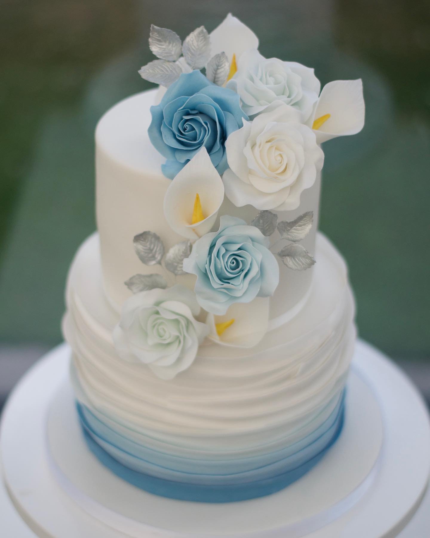 A two-tier wedding cake with a white top and blue ombre bottom, decorated with blue and white sugar flowers and leaves, elegantly displayed on a round base.