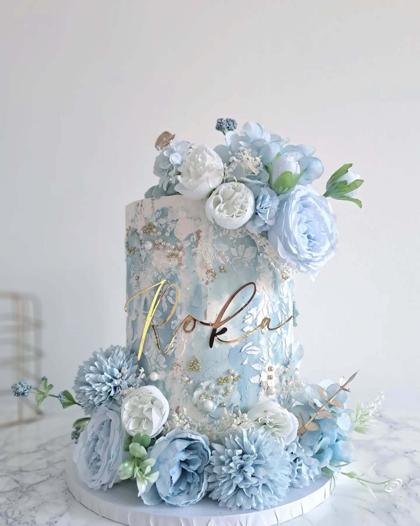 Elegant cake with blue and white flowers and gold script reading "Roka."