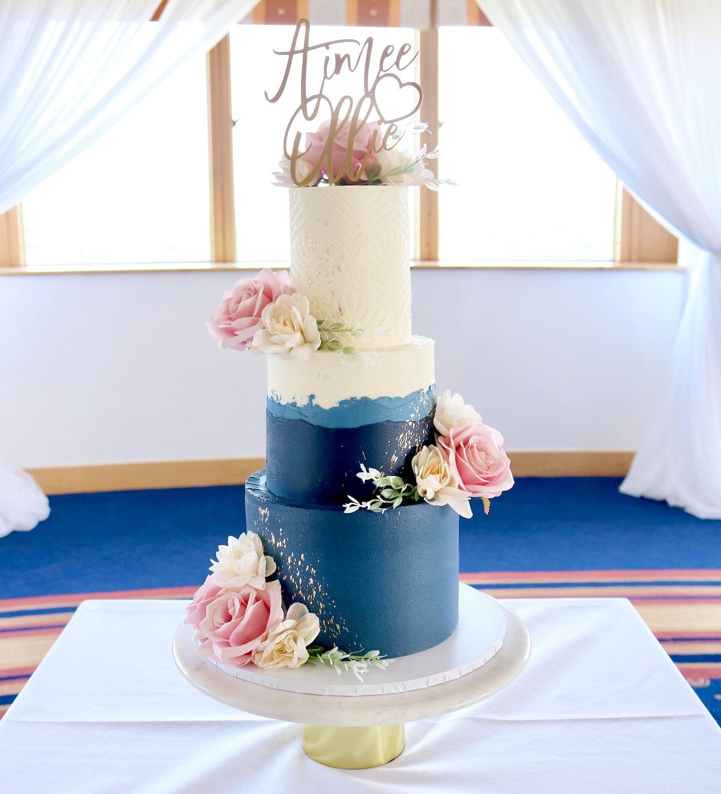 Three-tiered blue and white wedding cake with floral accents and personalized topper.