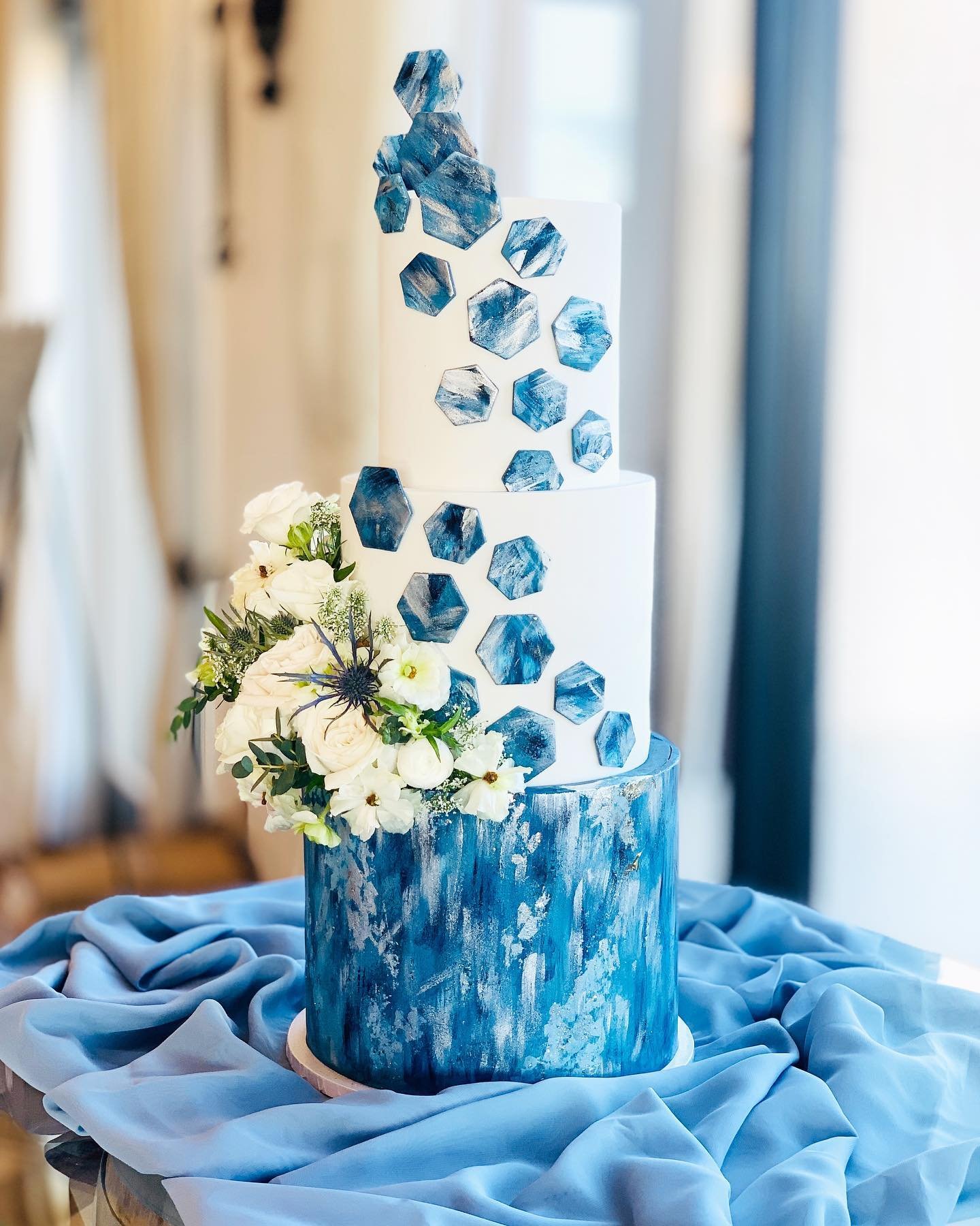 A three-tier wedding cake with white layers adorned with blue geode decorations and a marbled blue bottom, elegantly displayed on a table with soft fabric draping.