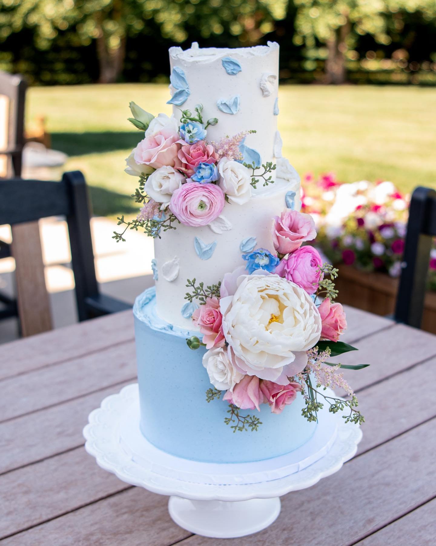 A three-tier wedding cake with a white top and soft blue bottom, decorated with blue petals and various flowers, elegantly displayed on a white pedestal in a garden setting.