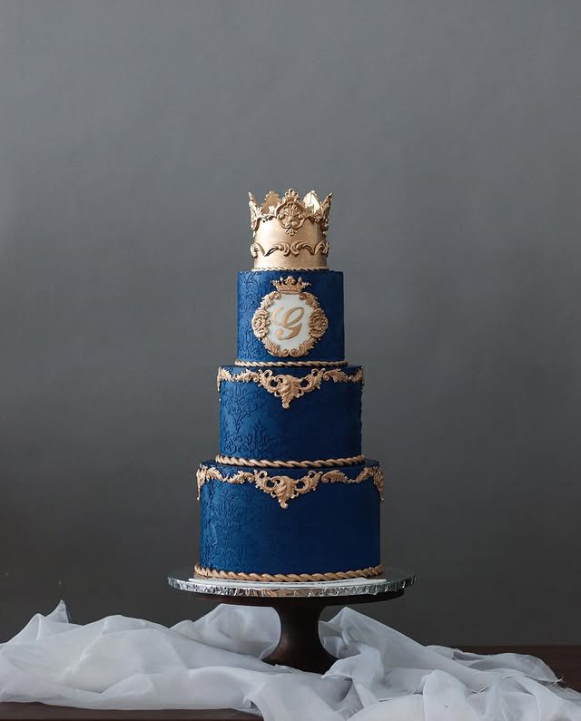 A four-tier cake with navy blue and gold, featuring a crown and lace decorations.