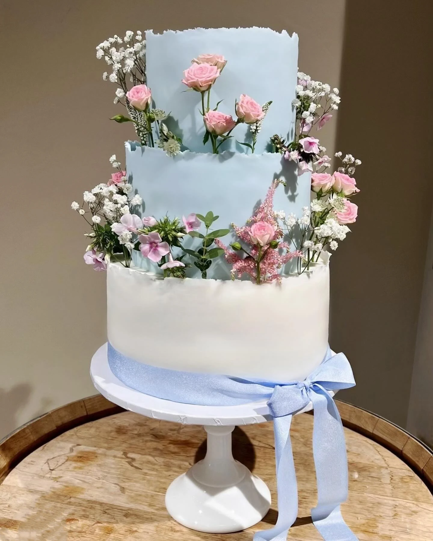 Three-tiered cake with a blue finish adorned with fresh flowers and a blue ribbon.