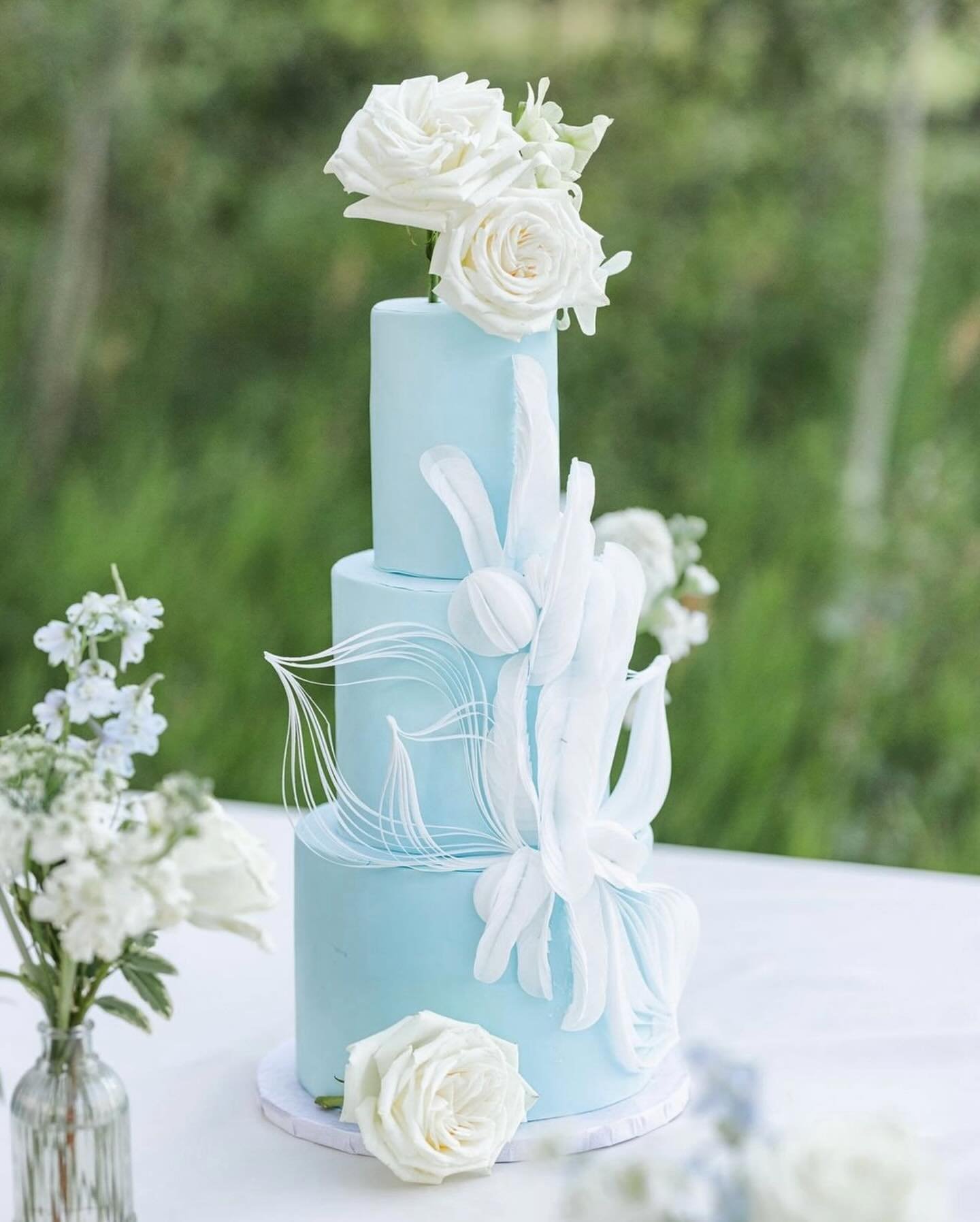 A blue three-tier cake with white floral decorations and white roses on top.