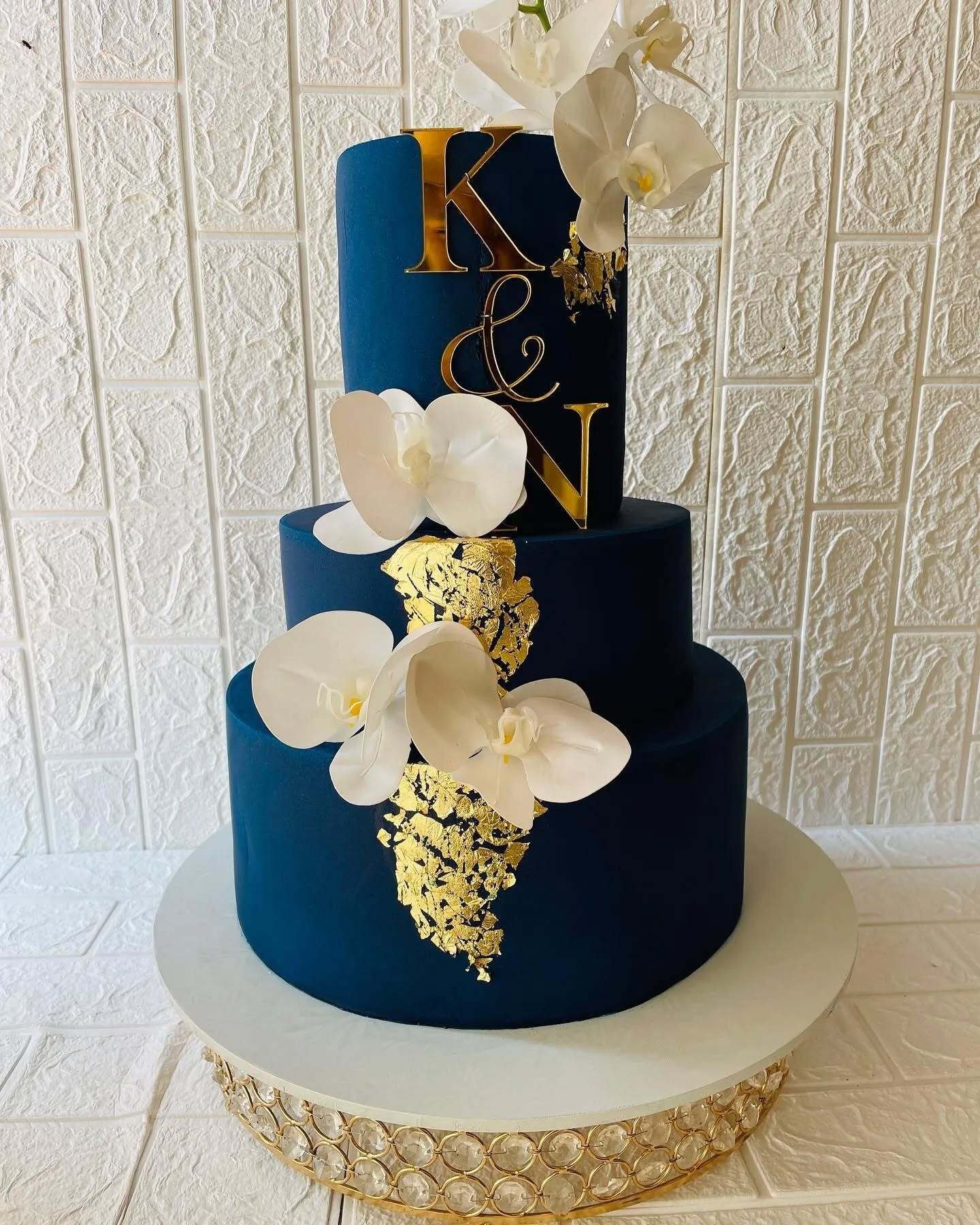 A three-tier cake with navy blue and gold, decorated with white orchids and initials.