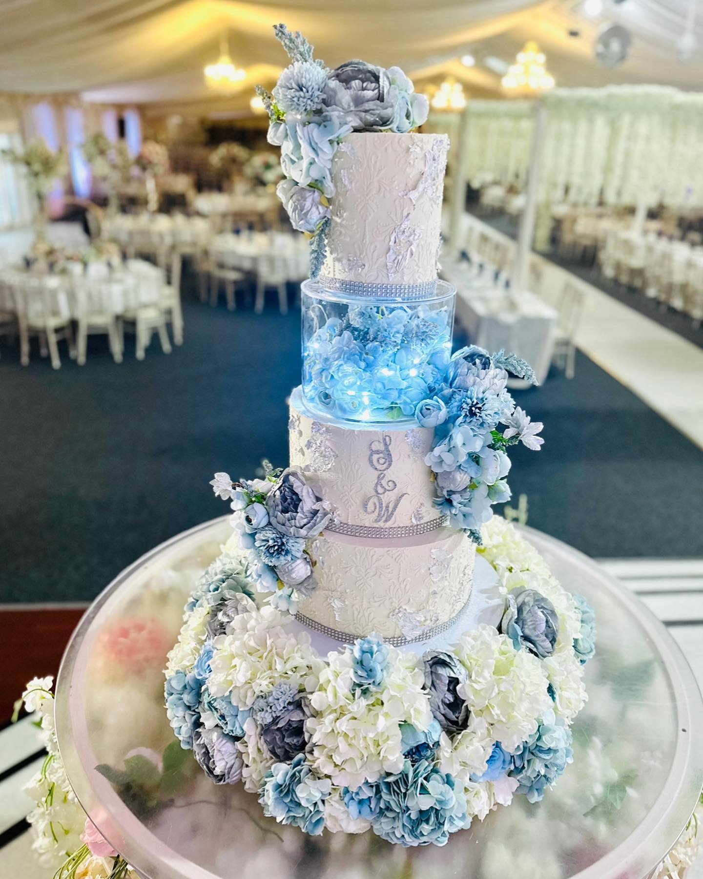 Four-tiered wedding cake with lace detailing, illuminated elements, and blue floral accents.