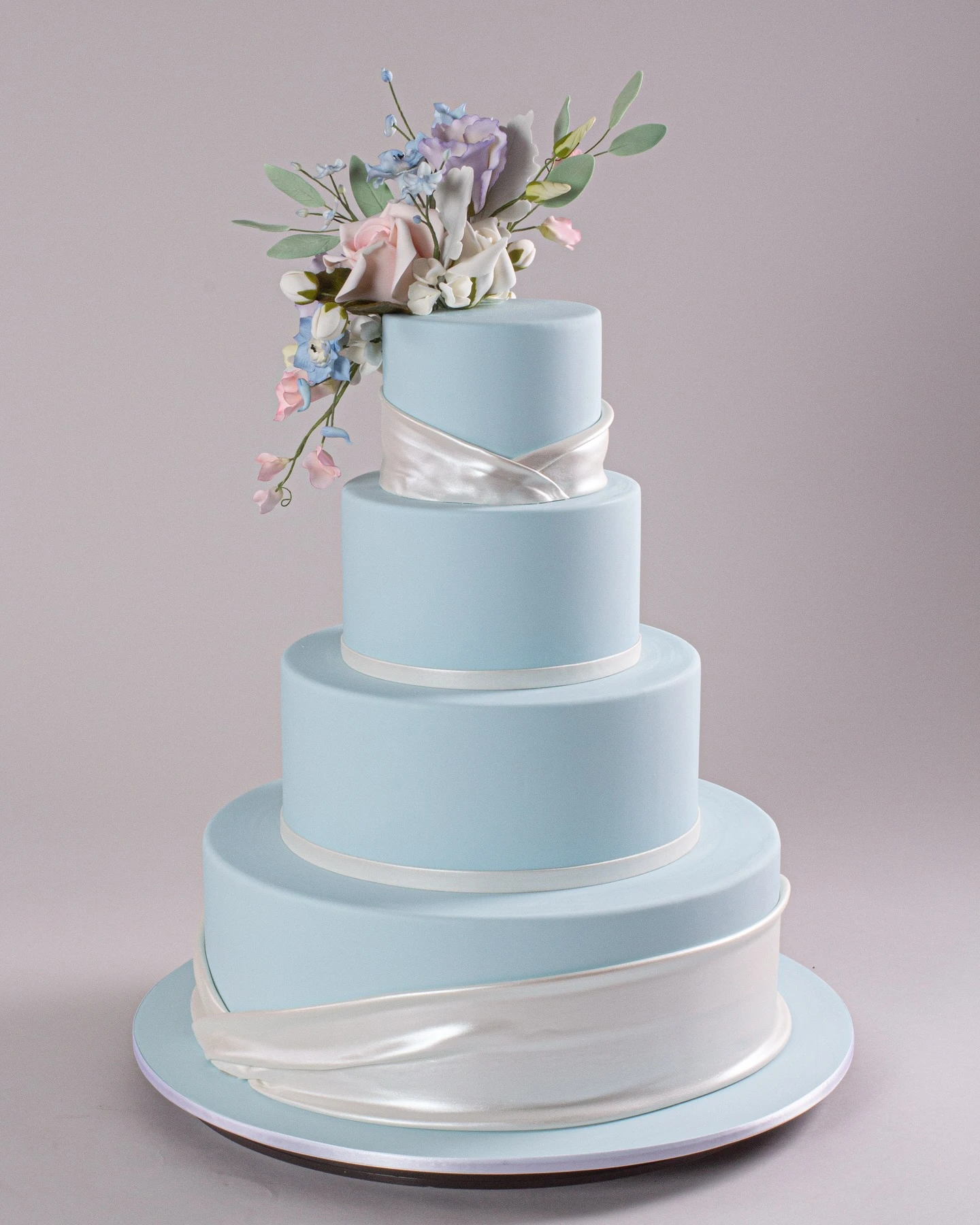 Four-tiered pastel blue cake adorned with a satin ribbon and sugar flowers on top.