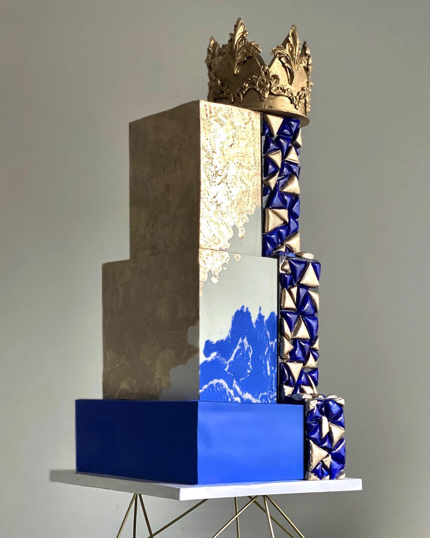 A three-tier cake with gold and blue, featuring geometric designs and a crown.