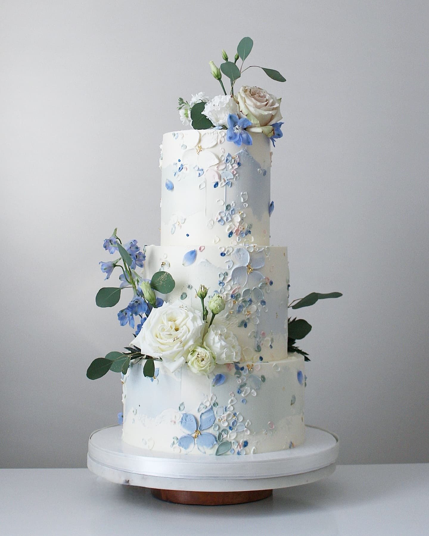 Three-tiered white cake decorated with edible flowers and greenery in soft colors.