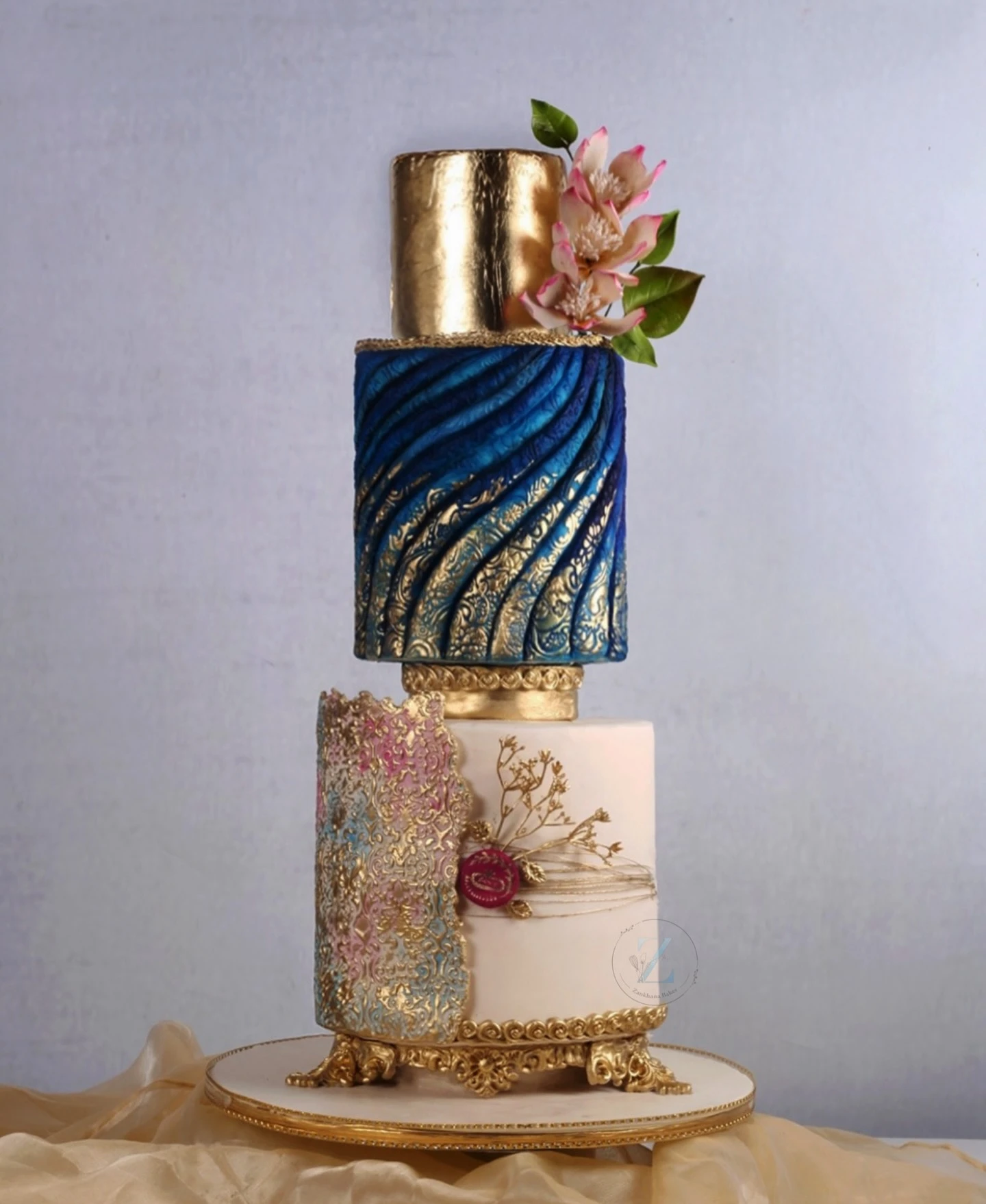 A three-tier cake with gold, blue, and pink, featuring textured designs and floral accents.