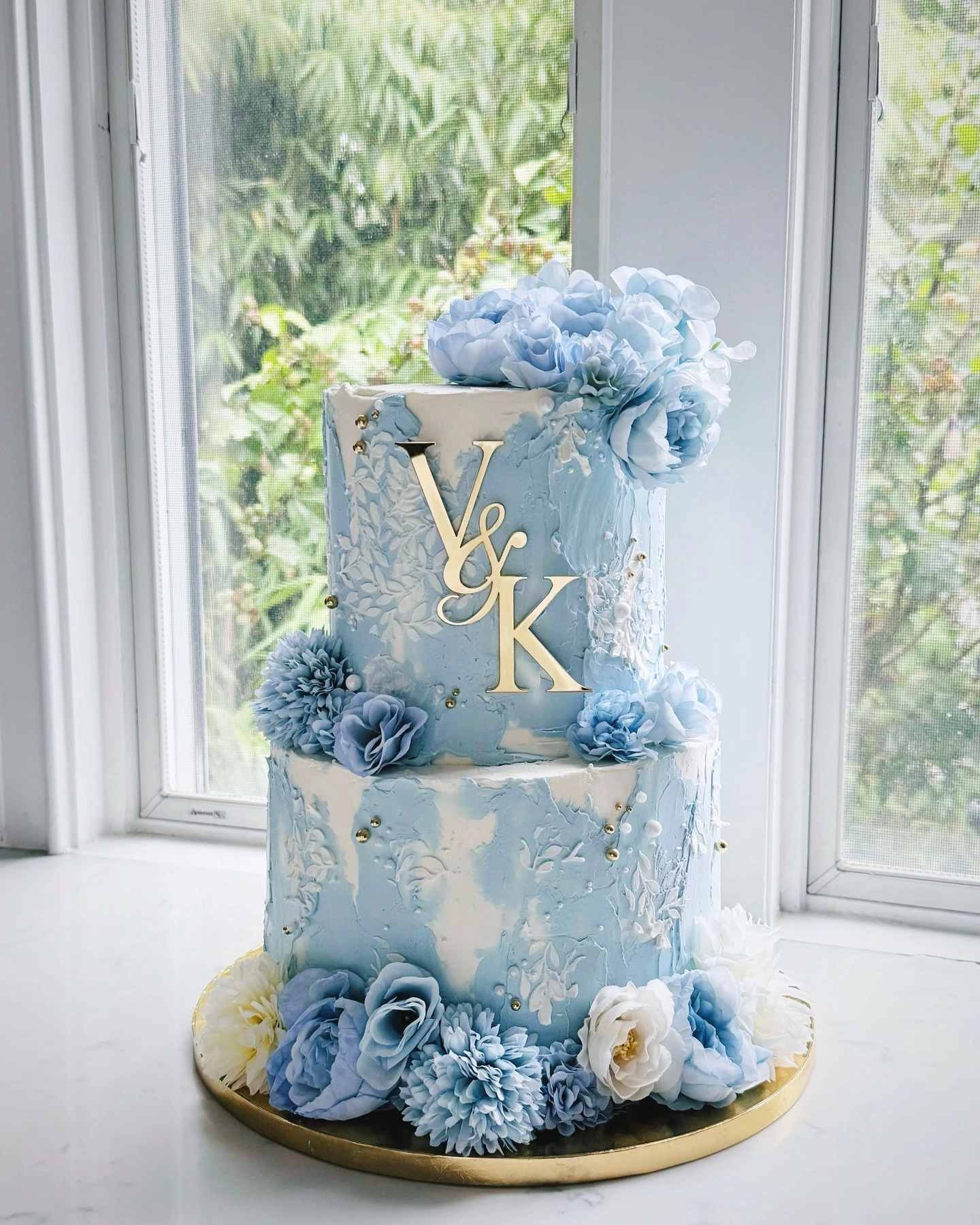 Two-tiered wedding cake adorned with blue flowers, gold accents, and a personalized monogram.