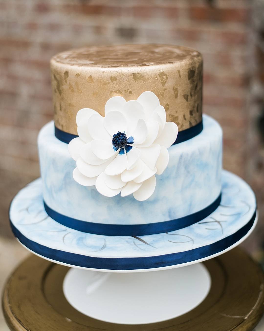 A two-tier cake with gold and blue, adorned with a white flower and navy ribbon.