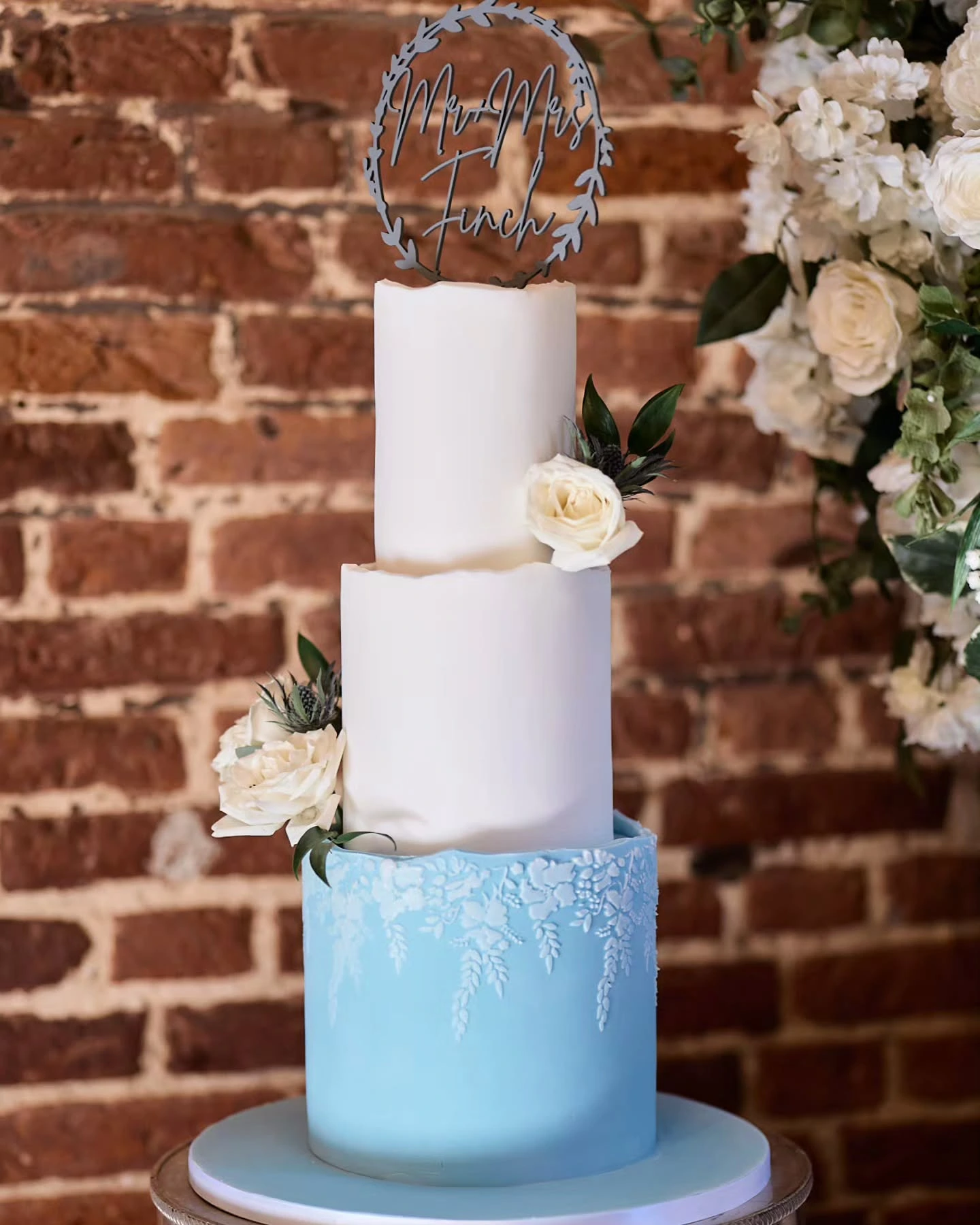 Three-tiered wedding cake with white and blue tiers, adorned with flowers and a personalized topper.