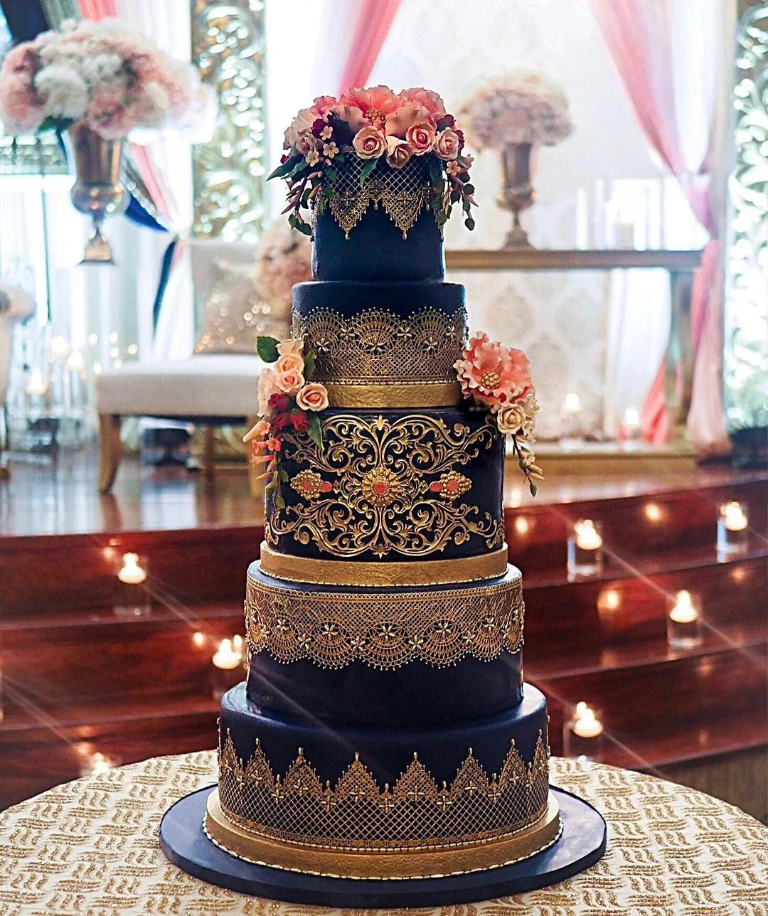 A five-tier wedding cake with navy blue layers, gold lace details, and floral accents.