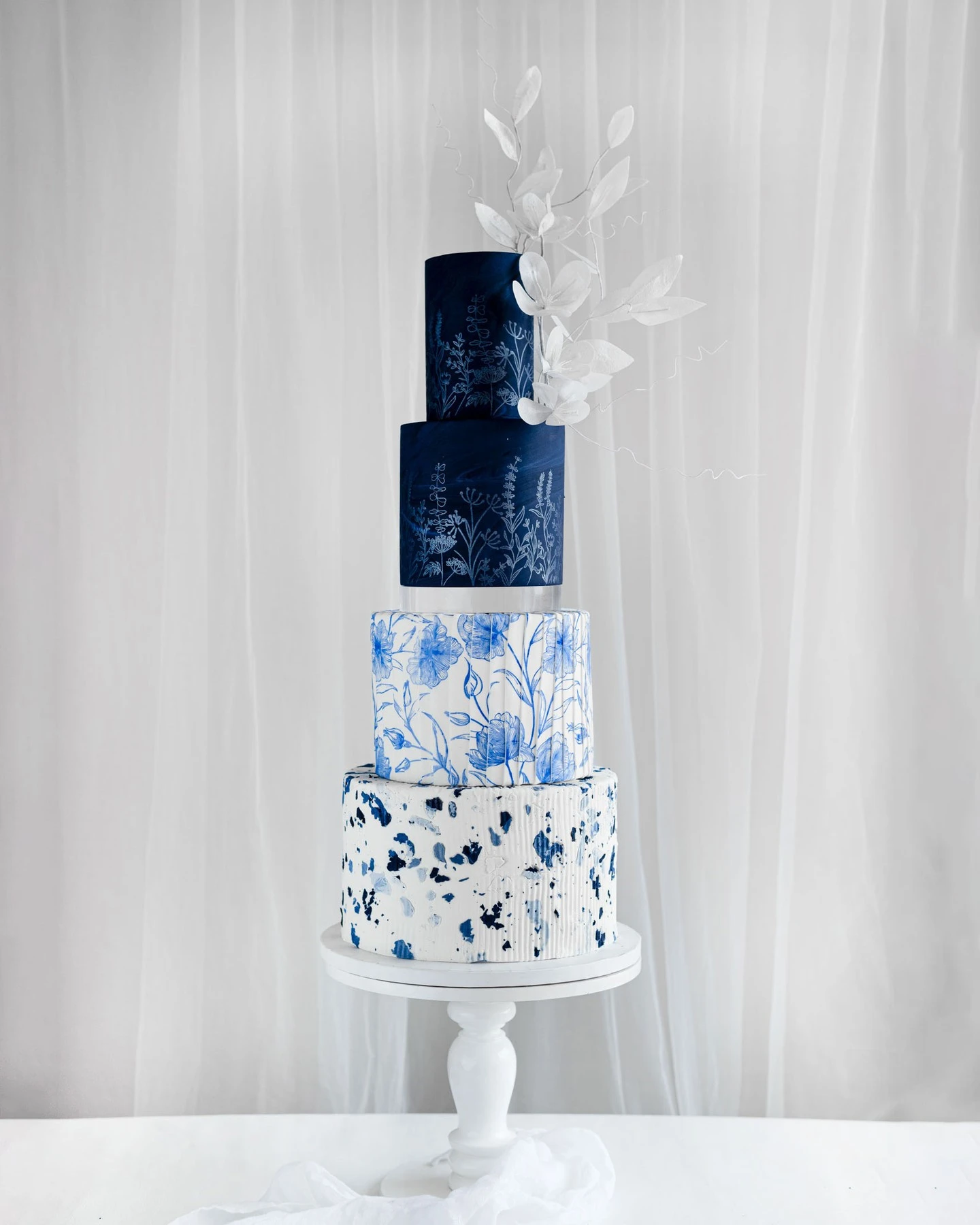 Four-tiered blue and white wedding cake with floral illustrations, abstract patterns, and white sugar leaves on a pedestal stand.