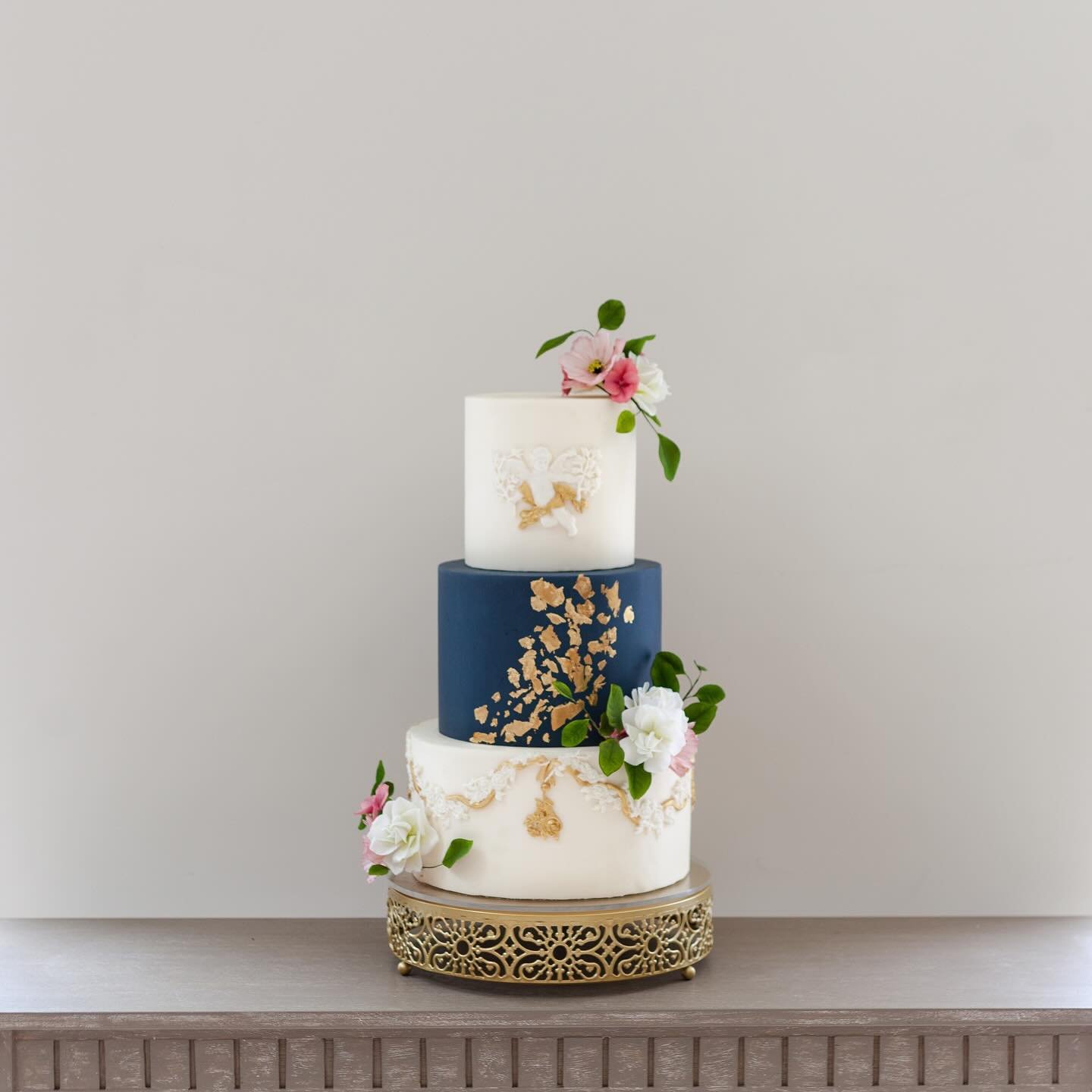 A three-tier wedding cake with a blue middle layer and white top and bottom, decorated with flowers and gold accents, displayed on a decorative stand against a neutral background.