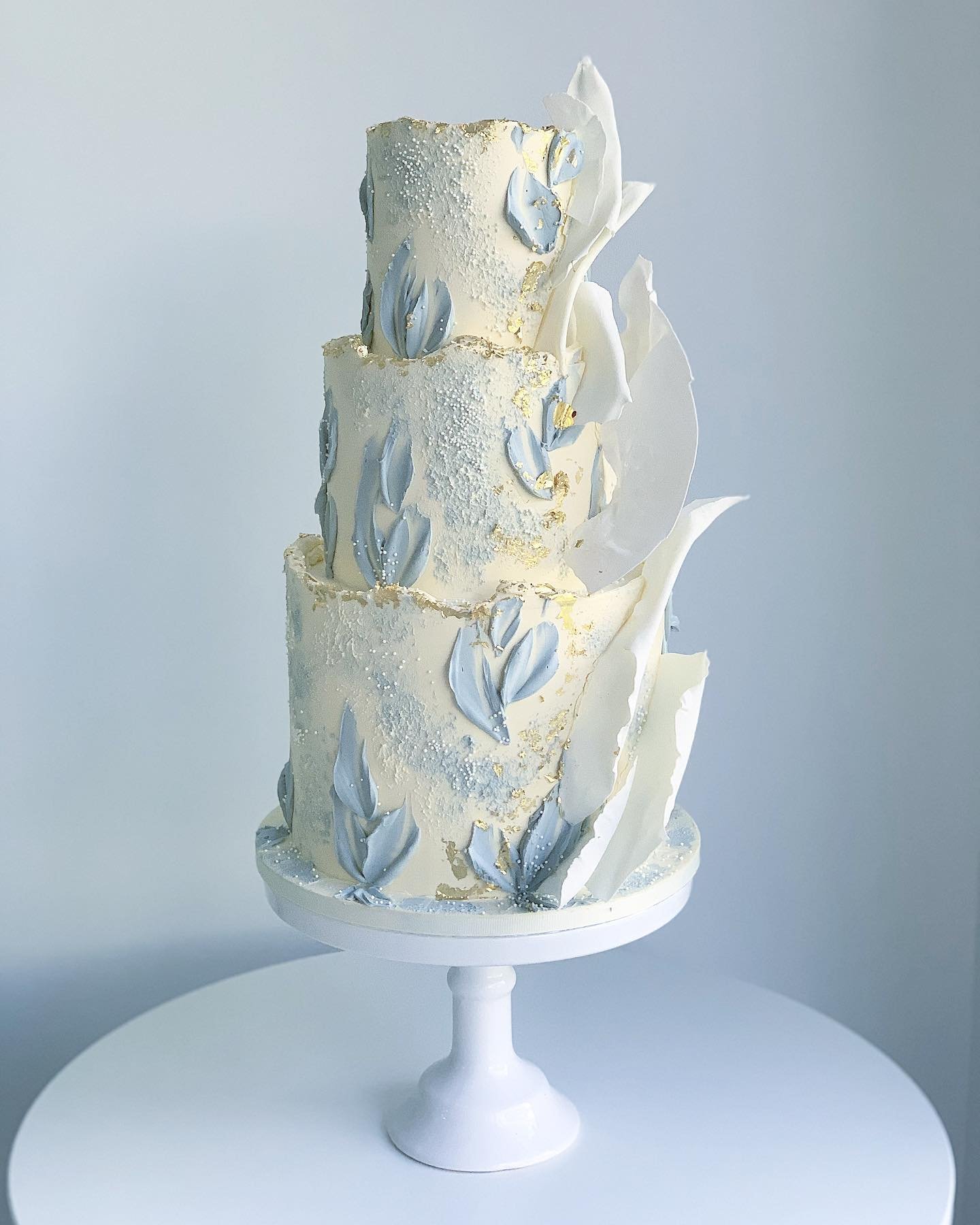 Three-tier light blue wedding cake with white petals and gold accents.