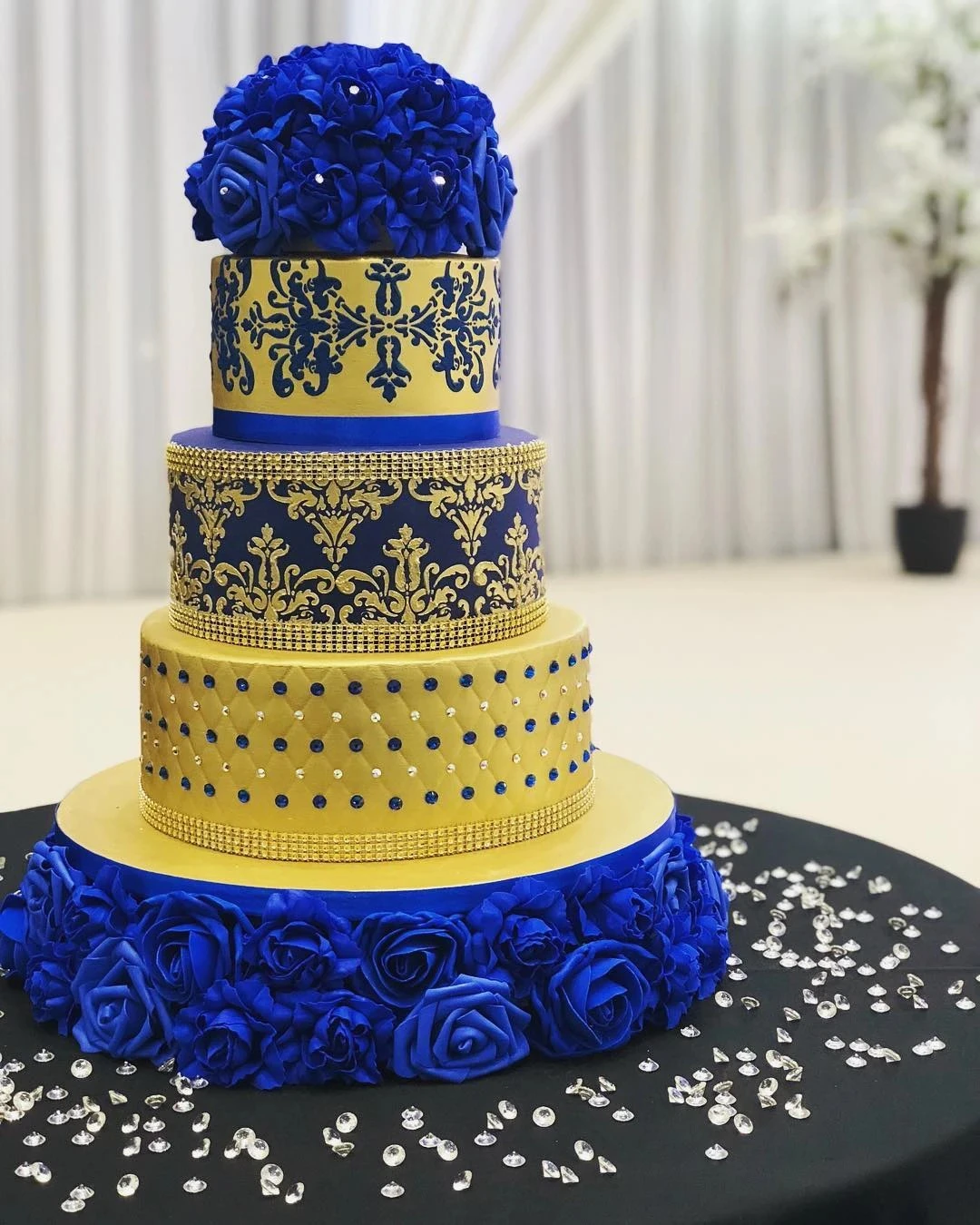 A four-tier cake with blue and gold designs, adorned with blue roses and rhinestones.