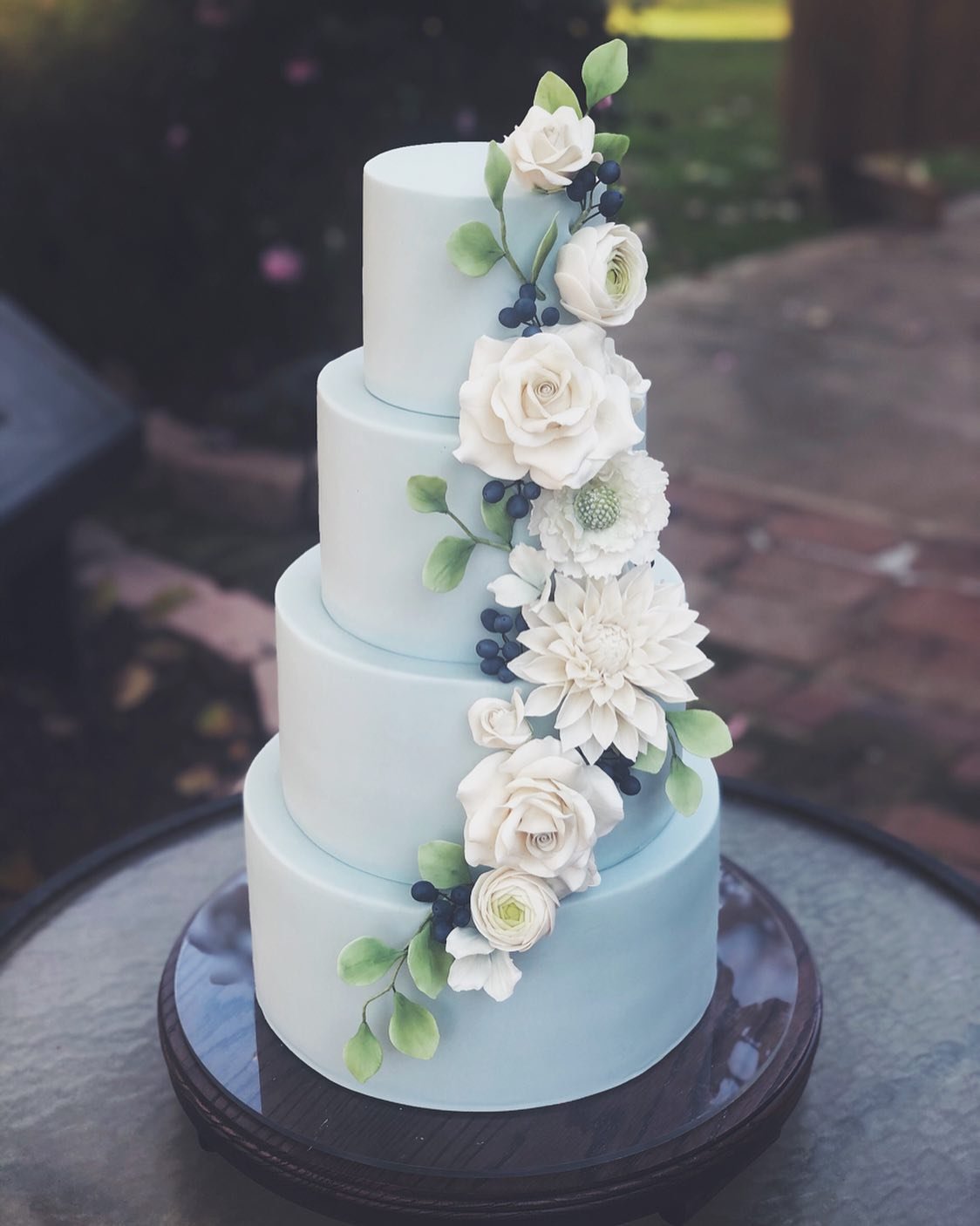 Four-tiered light blue cake with white flowers and green leaves.