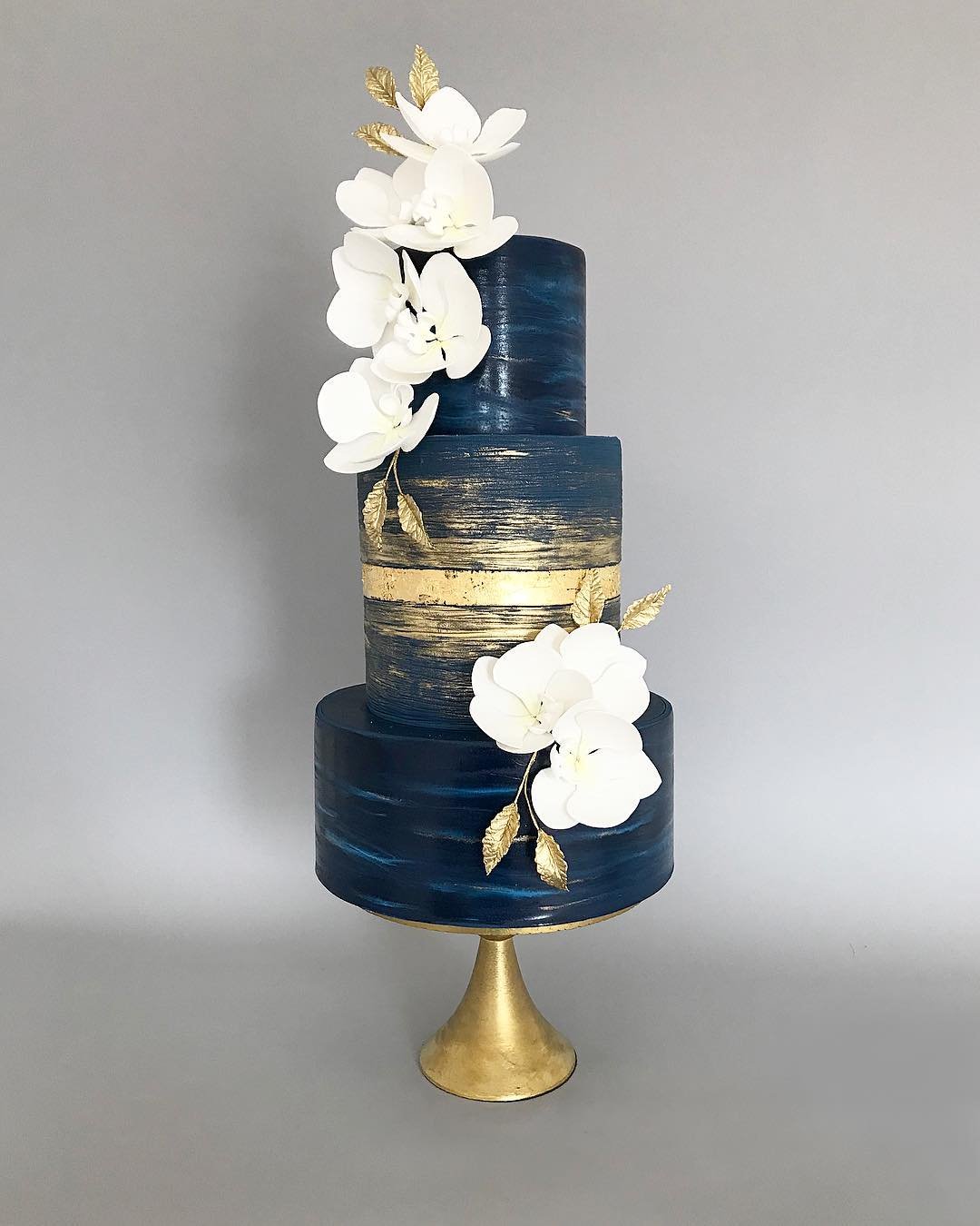 A three-tier cake with navy blue layers, gold accents, and white orchid decorations.