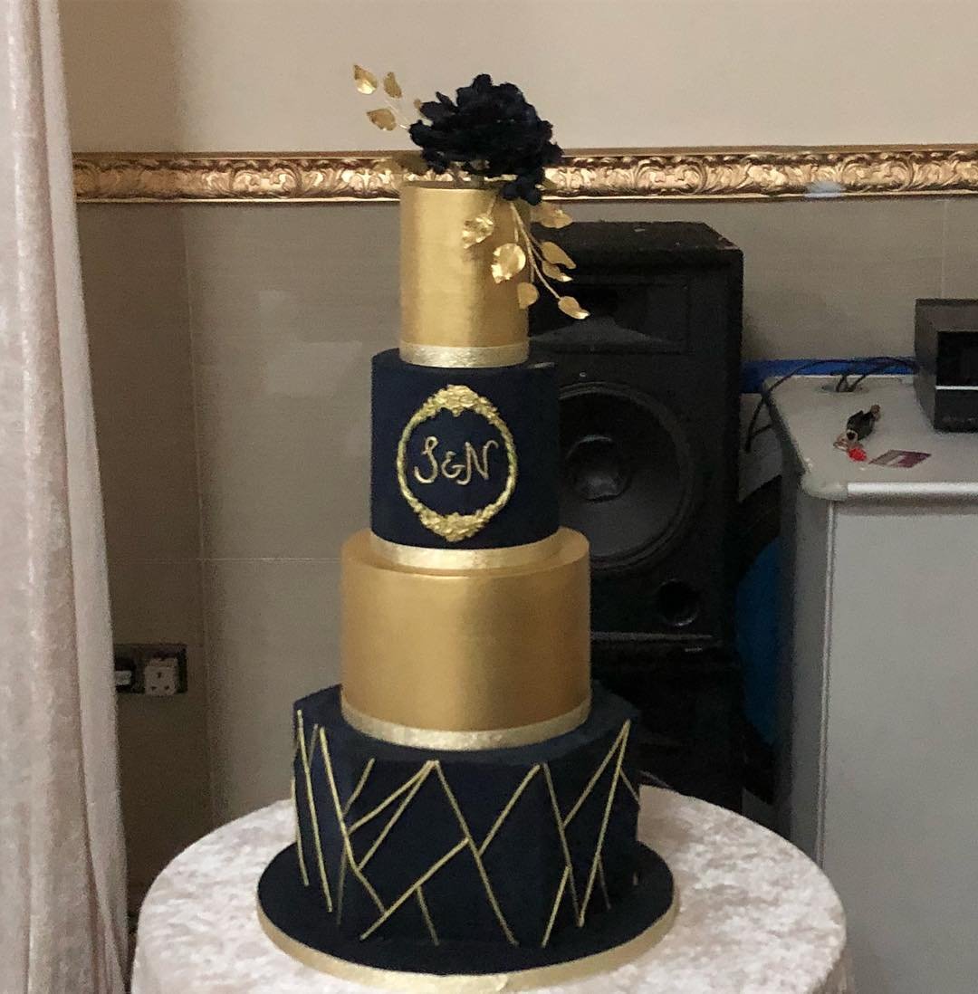 A four-tier cake with navy blue and gold designs, topped with a black flower and Golden leaves.