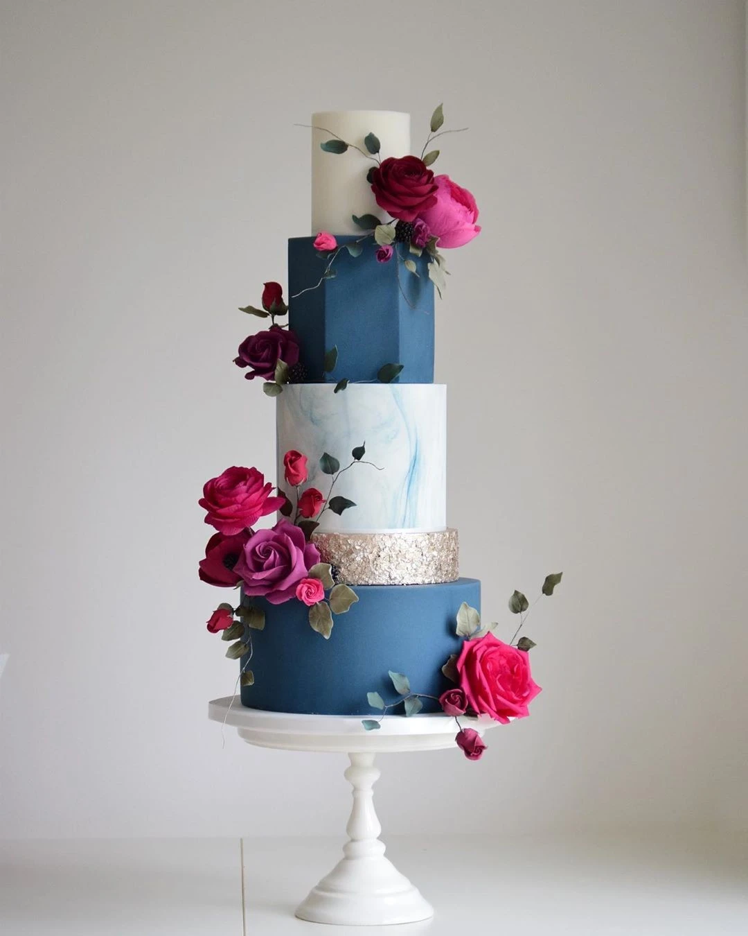 A four-tier wedding cake with deep blue and marbled white layers, decorated with pink roses and greenery, featuring a gold accent band.