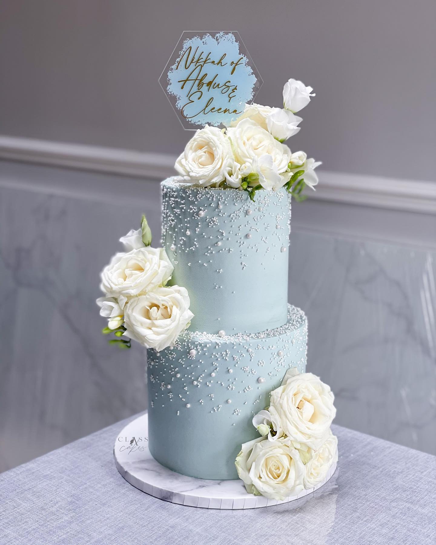 Two-tiered blue wedding cake decorated with white roses and edible pearls, featuring a personalized cake topper.