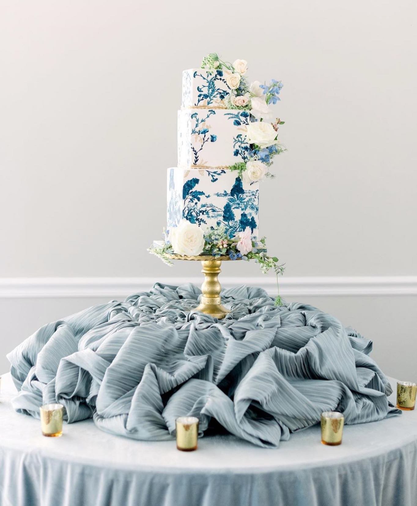 A three-tier wedding cake with blue floral designs on a white surface, decorated with fresh flowers and greenery, displayed on a golden stand.