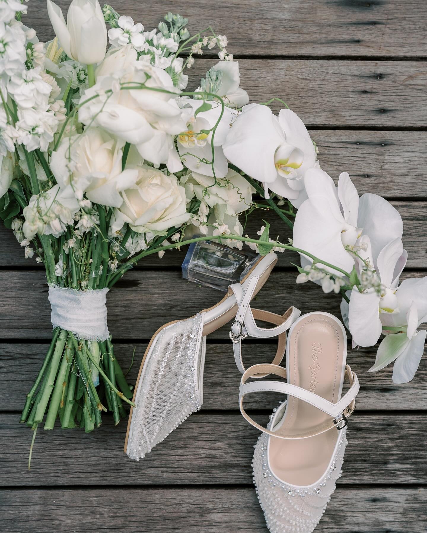 A white bridal bouquet featuring roses, orchids, and other white flowers, next to wedding shoes.