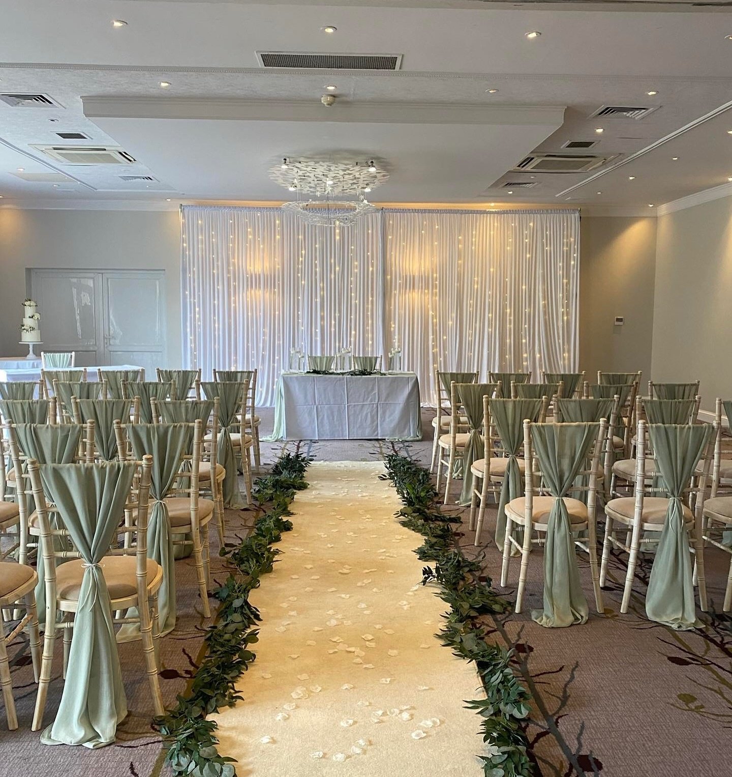 A wedding ceremony setup with rows of chairs draped in sage green fabric, flanking a white aisle runner decorated with green foliage garlands.