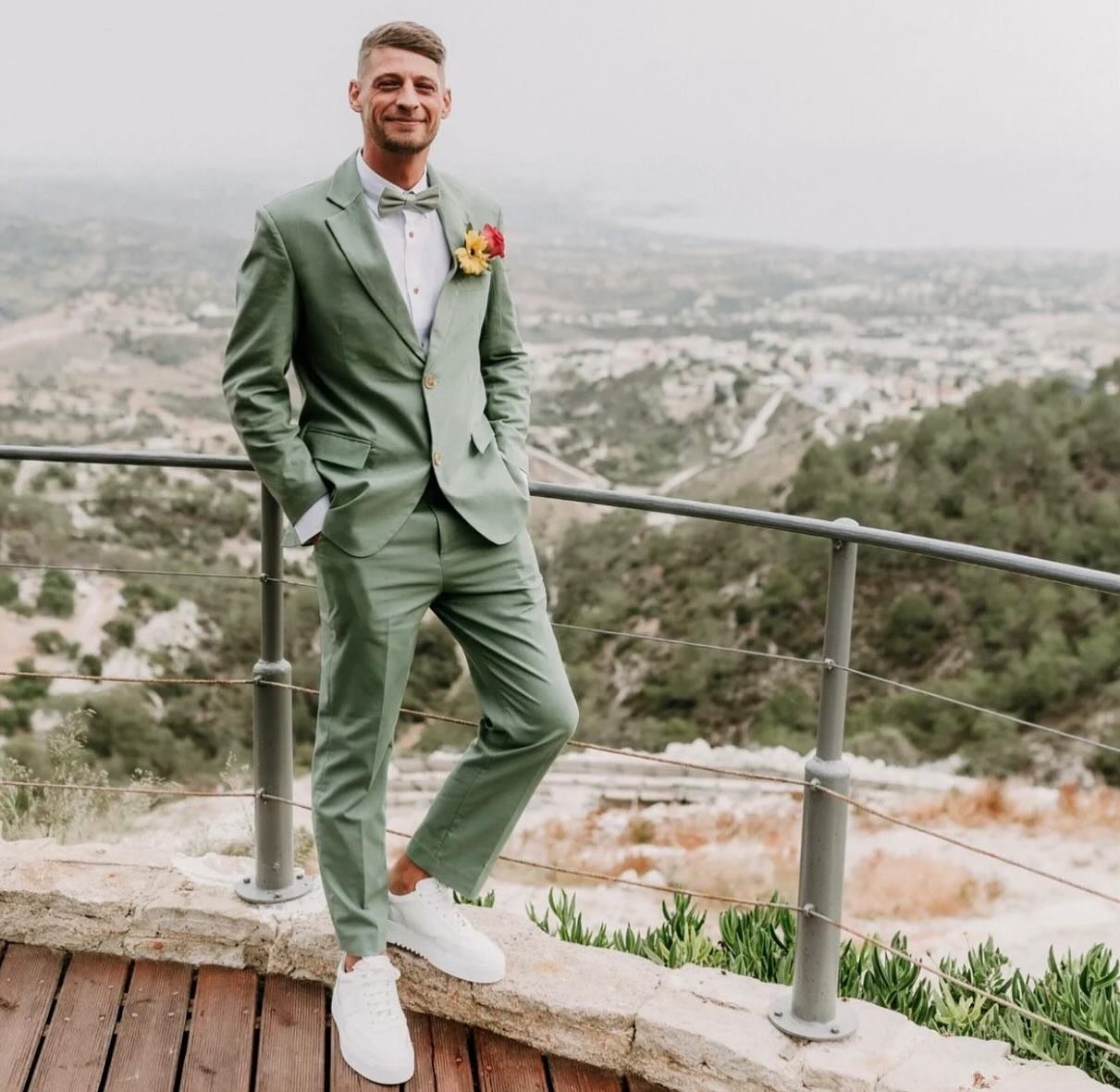 A groom smiling and standing outdoors, wearing a sage green suit, a white shirt, a light bow tie, white sneakers, and a colorful boutonnière.