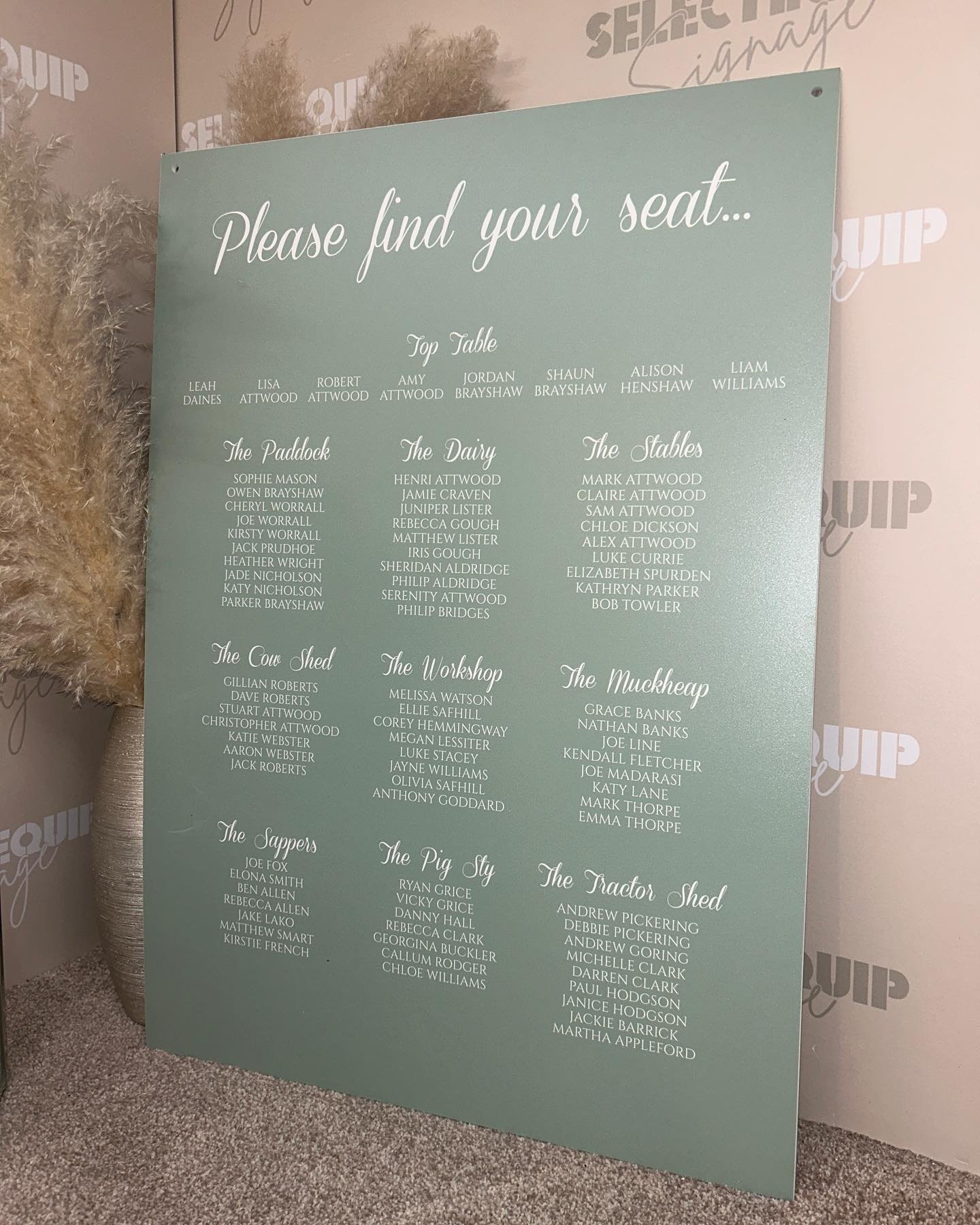 A tall, clear or frosted acrylic wedding seating chart with guest names listed under table names, featuring a sage green color element in its design.