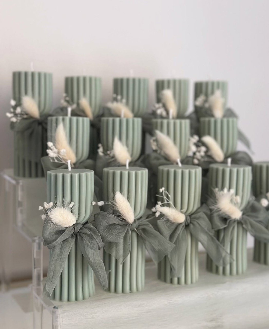 Multiple tall, ridged pillar candles in a soft sage green color, each tied with a sheer sage green ribbon bow, arranged together on a clear surface.